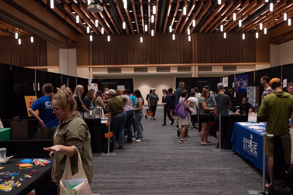 Indoor event with people interacting at various booths under a ceiling with hanging lights.
