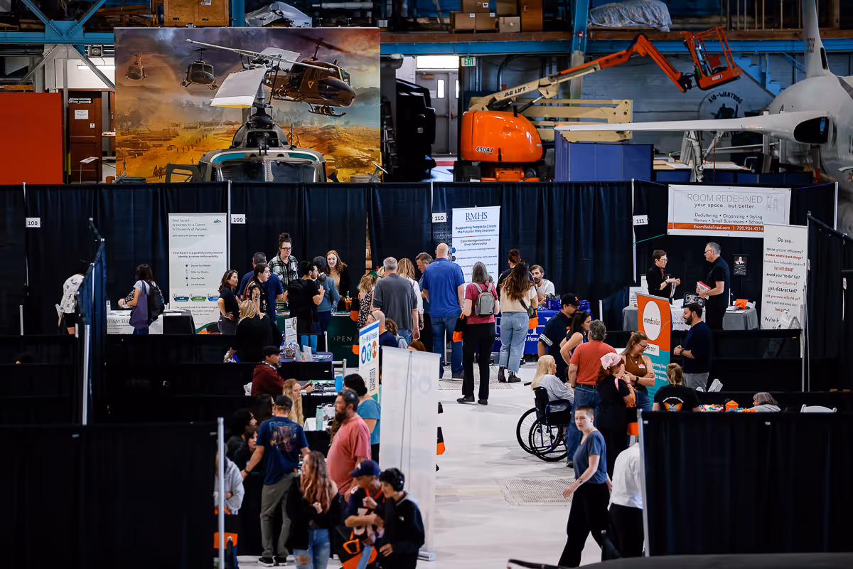 Indoor event with multiple booths and attendees engaging in conversations, with an aircraft and helicopter display in the background.
