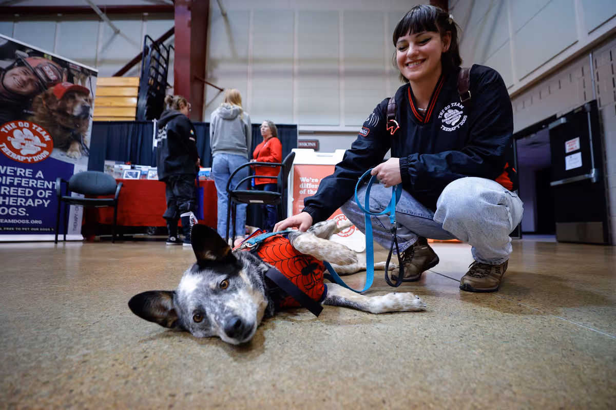 Smiling woman petting a lying dog wearing a red Spider-Man vest inside an event hall.