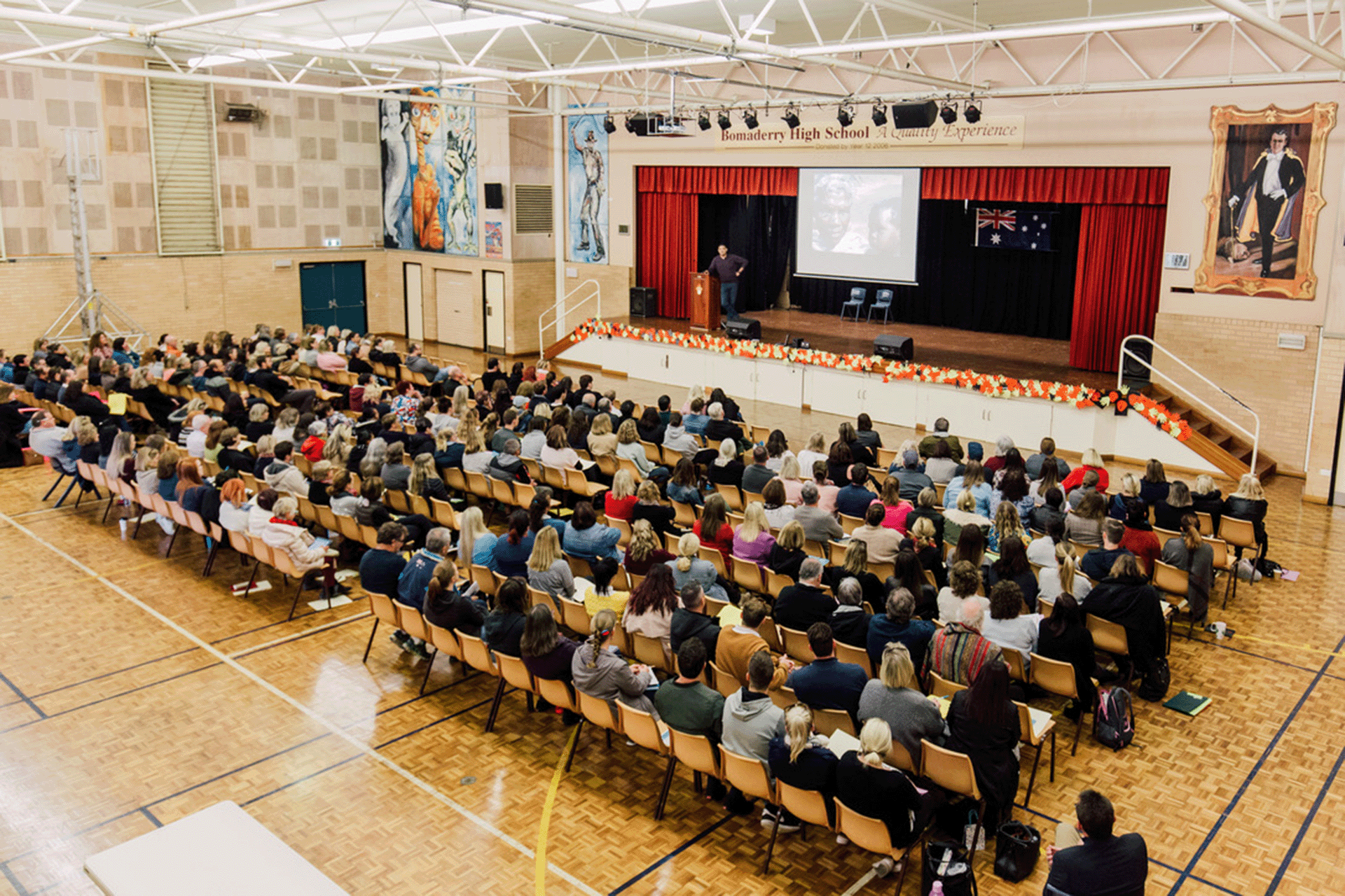 Monty Badami delivering a keynote on cultural intelligence and human connection to a large audience in a school auditorium, with visual storytelling and inclusive leadership themes on display.