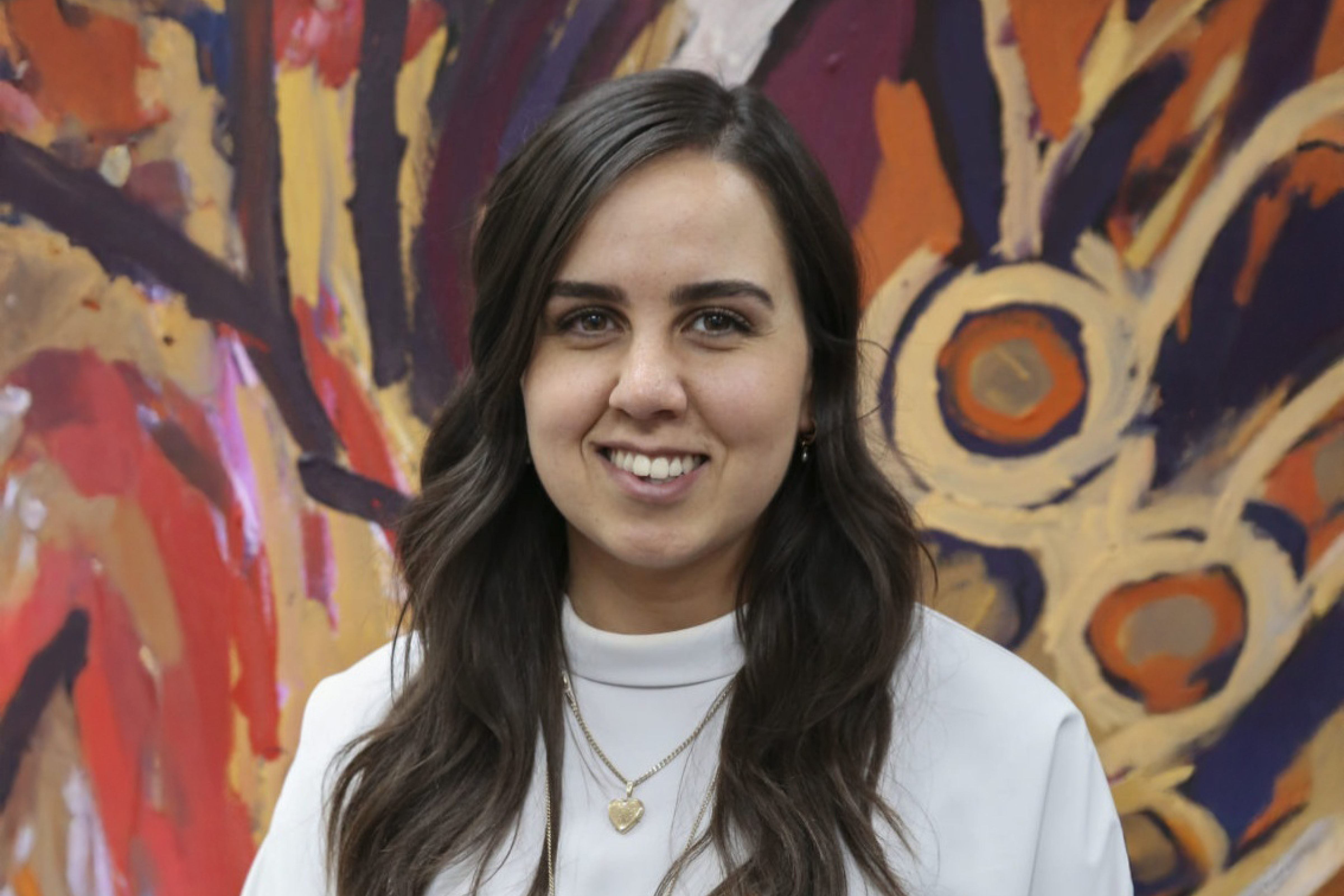 Smiling woman with long dark hair wearing a white top and layered necklaces, standing in front of a colorful abstract painting.
