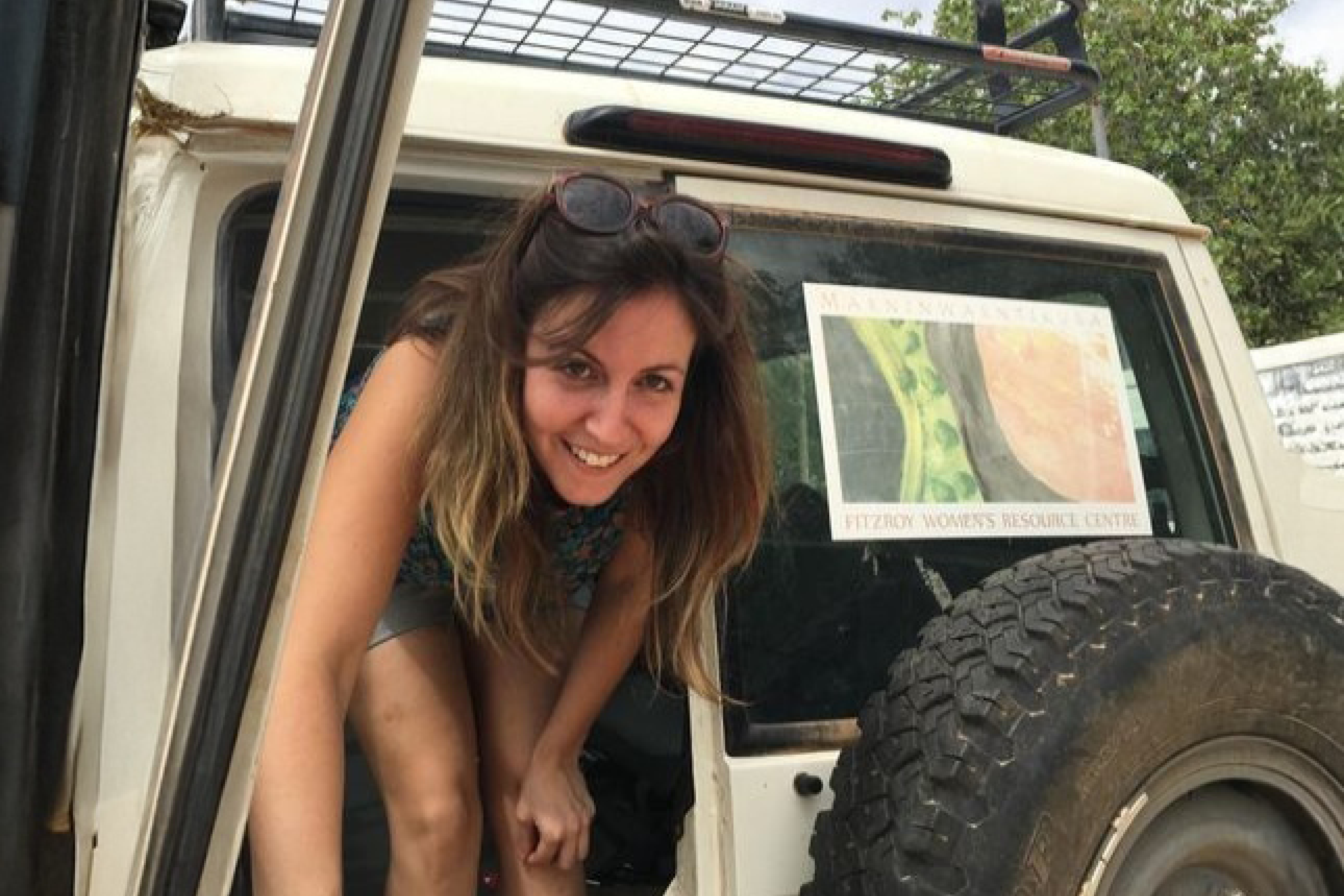Smiling woman with sunglasses on her head leaning out from the back of a white vehicle.