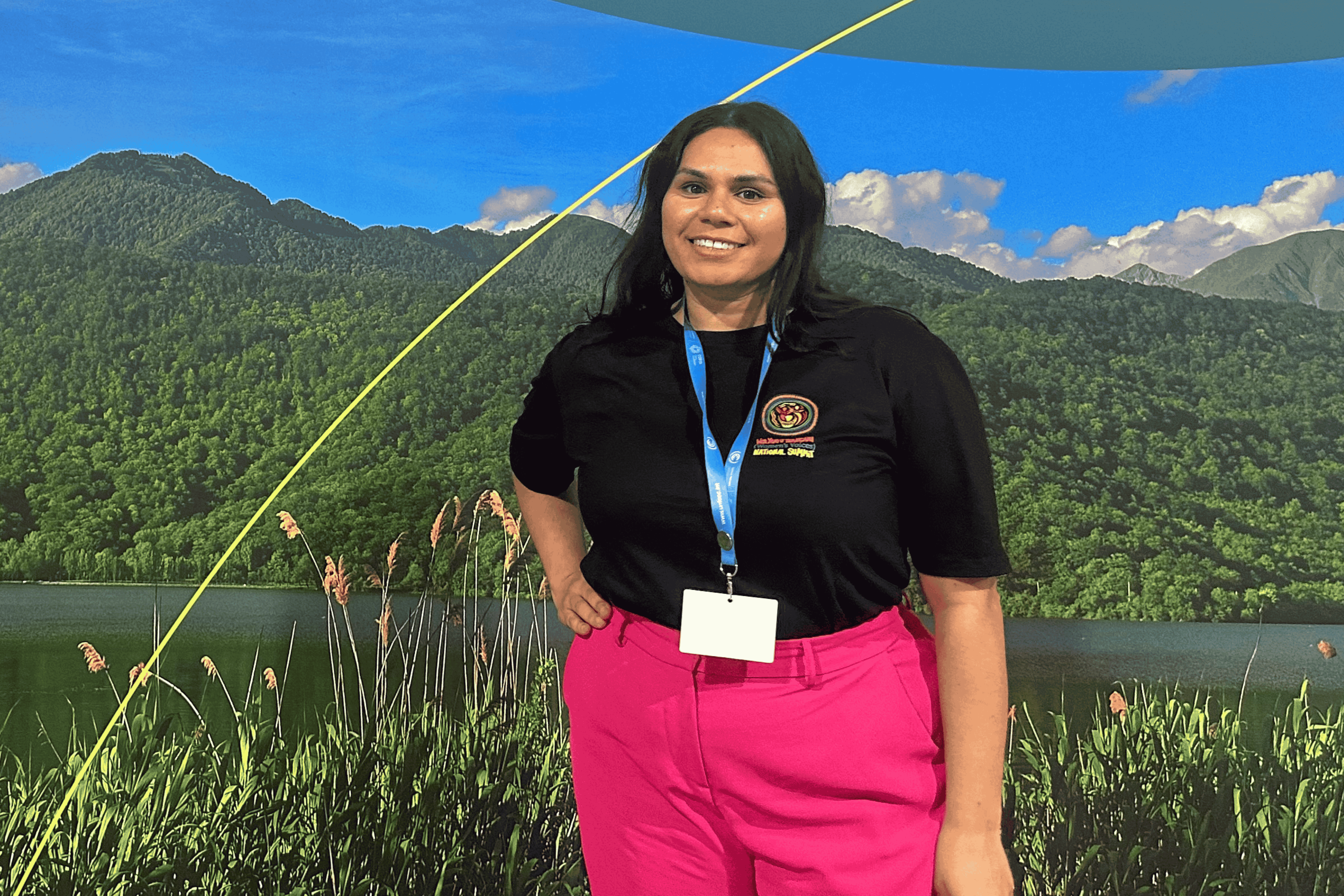 Woman with long dark hair wearing a black t-shirt and pink pants smiling in front of a scenic backdrop with green mountains and blue sky.