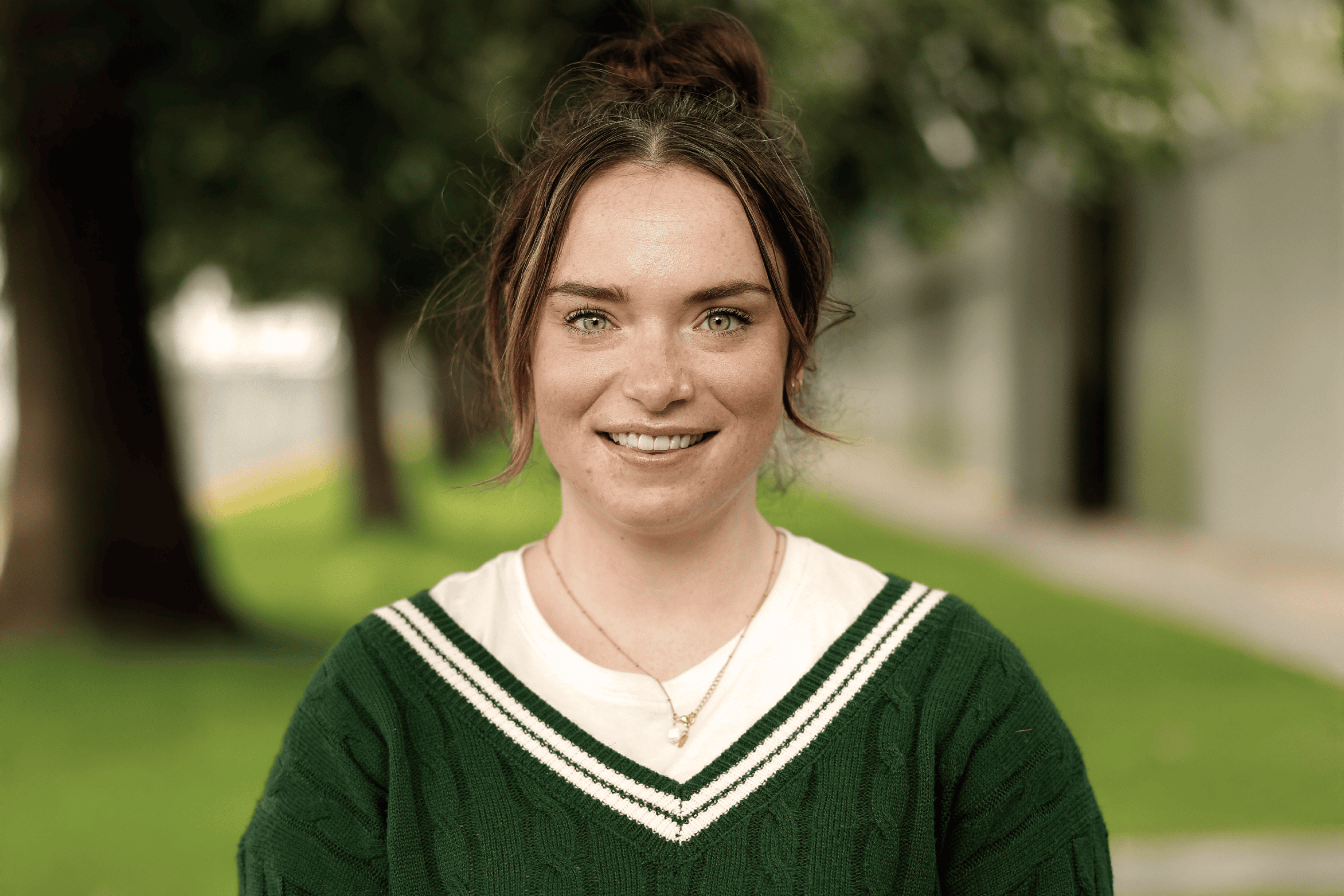 Smiling woman with brown hair tied up, wearing a green sweater with white stripes, standing outdoors with blurred greenery in the background.