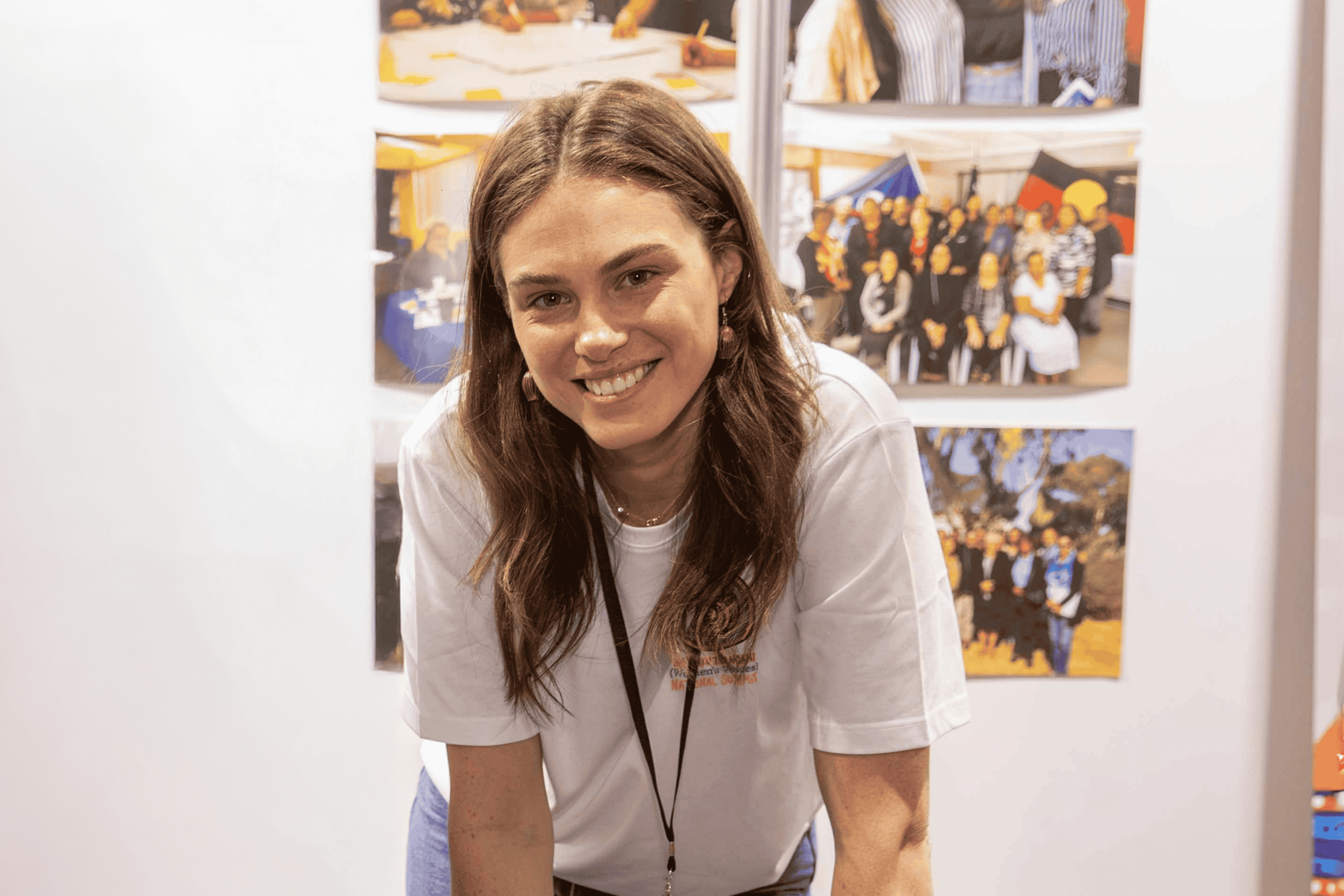 Young woman with long brown hair smiling, wearing a white t-shirt and a conference badge, leaning forward slightly.
