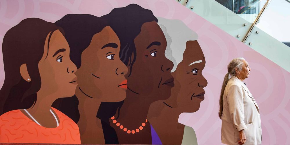 Photo of Aunty Matilda House at the Wiyi Yani U Thangani National Summit posing with a drawing of women on the wall. 