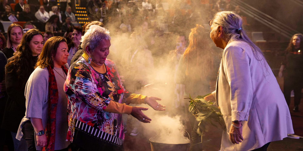 Photo of a smoking ceremony. People lining up to the smoking leaves with women placing her hands in the smoke and an Elder woman facing her