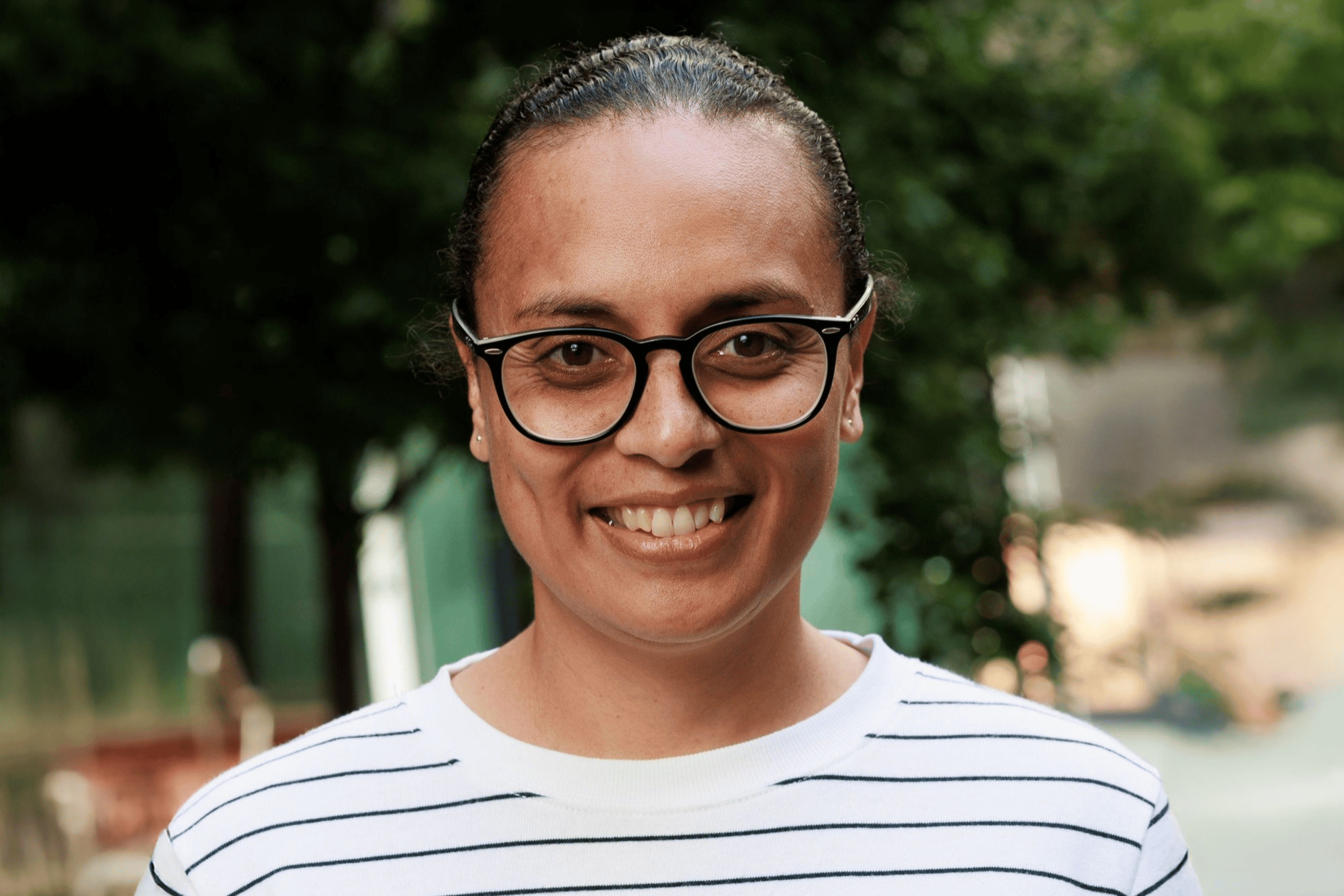 Smiling person with glasses and hair tied back wearing a white shirt with black horizontal stripes in an outdoor setting.