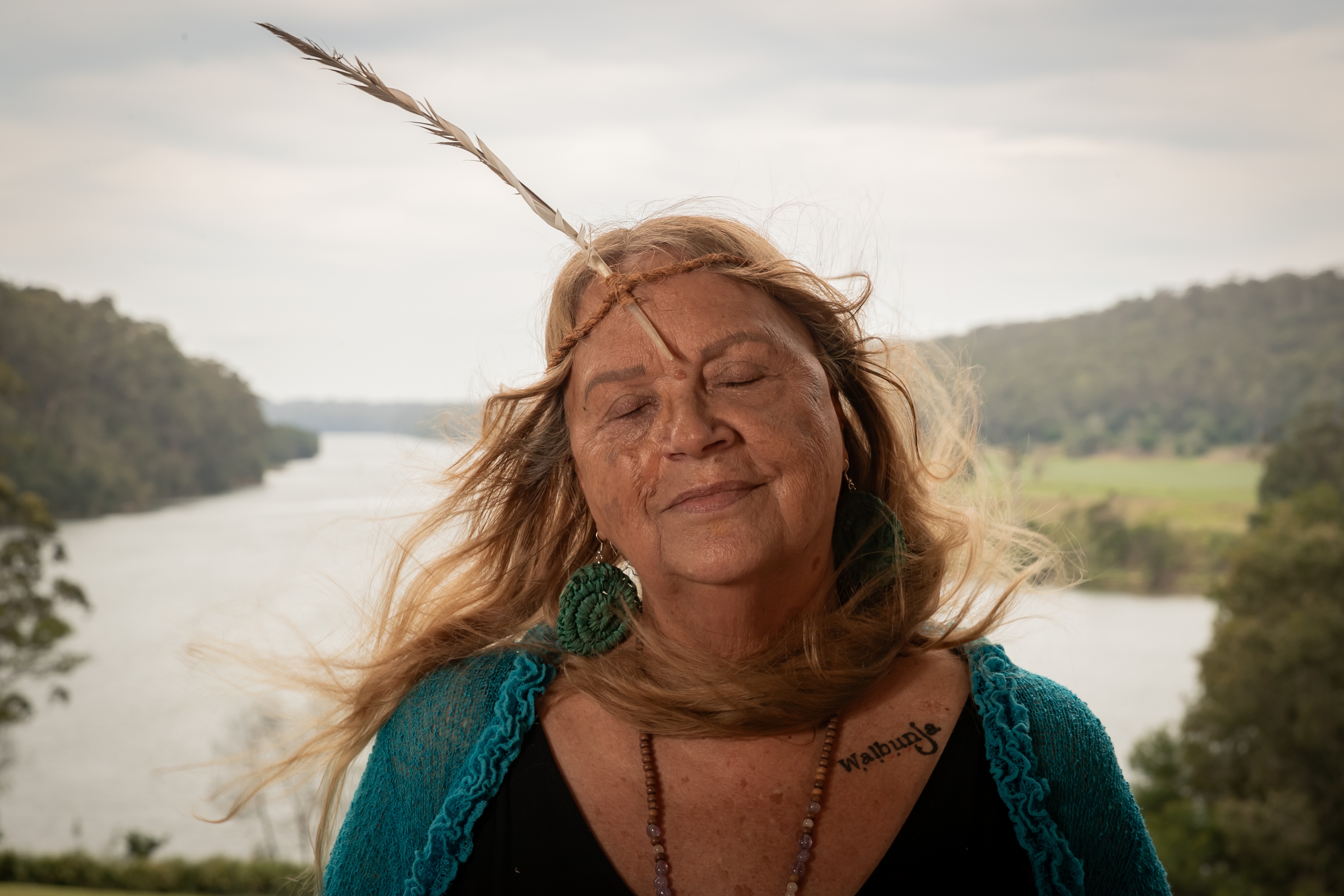 Older woman with long blonde hair, eyes closed, wearing a teal knitted shawl, large round earrings, necklace, and a feather headband, standing outdoors by a river with hills in the background.