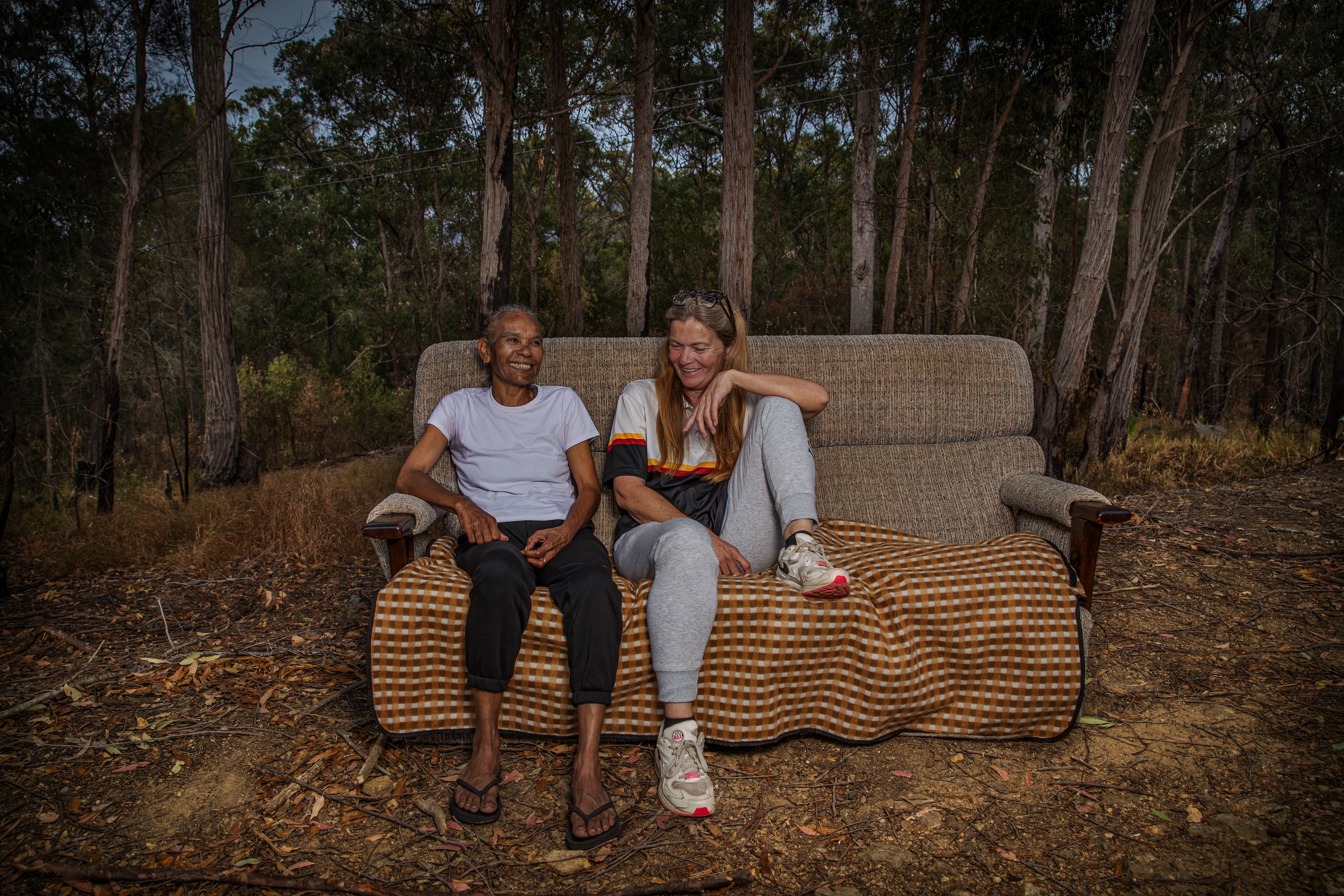 Two women sitting on a couch with a checkered blanket outdoors in a wooded area, smiling and engaging with each other.