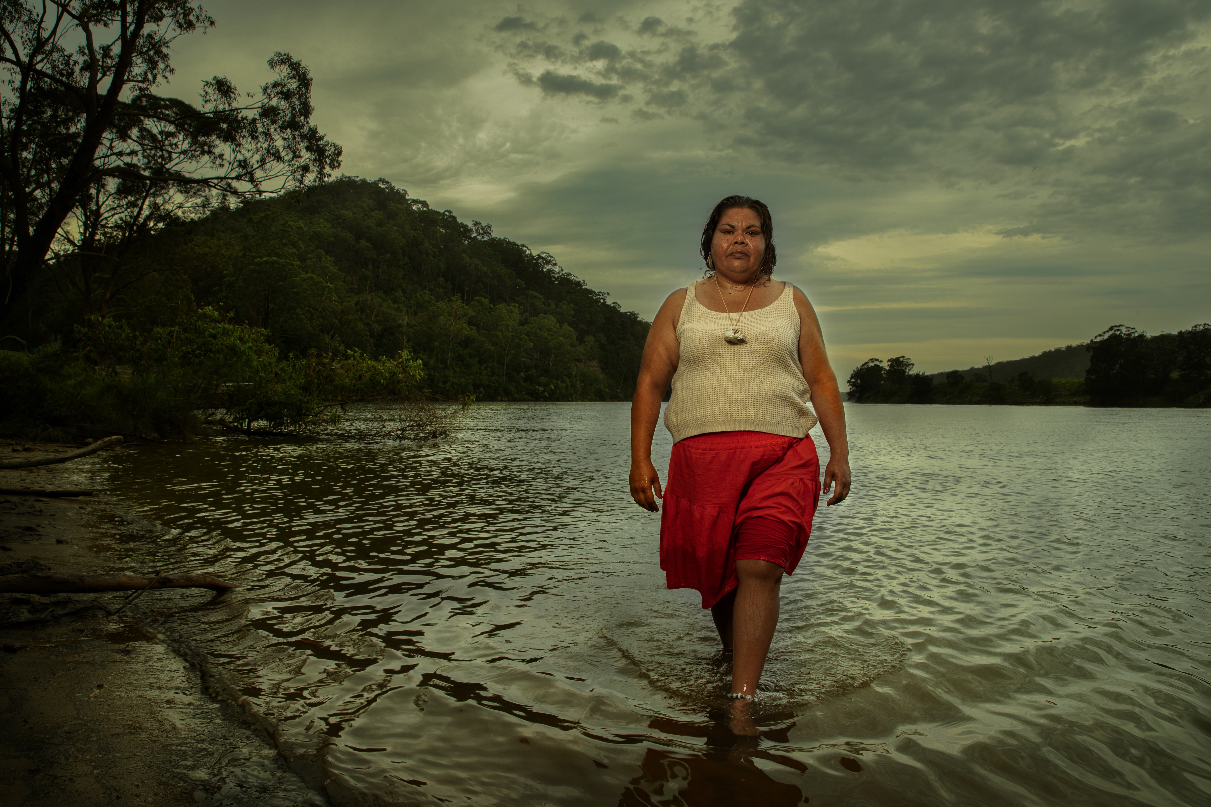 Woman in a white sleeveless top and red skirt walking through shallow water of a river with forested hills and cloudy sky in the background.