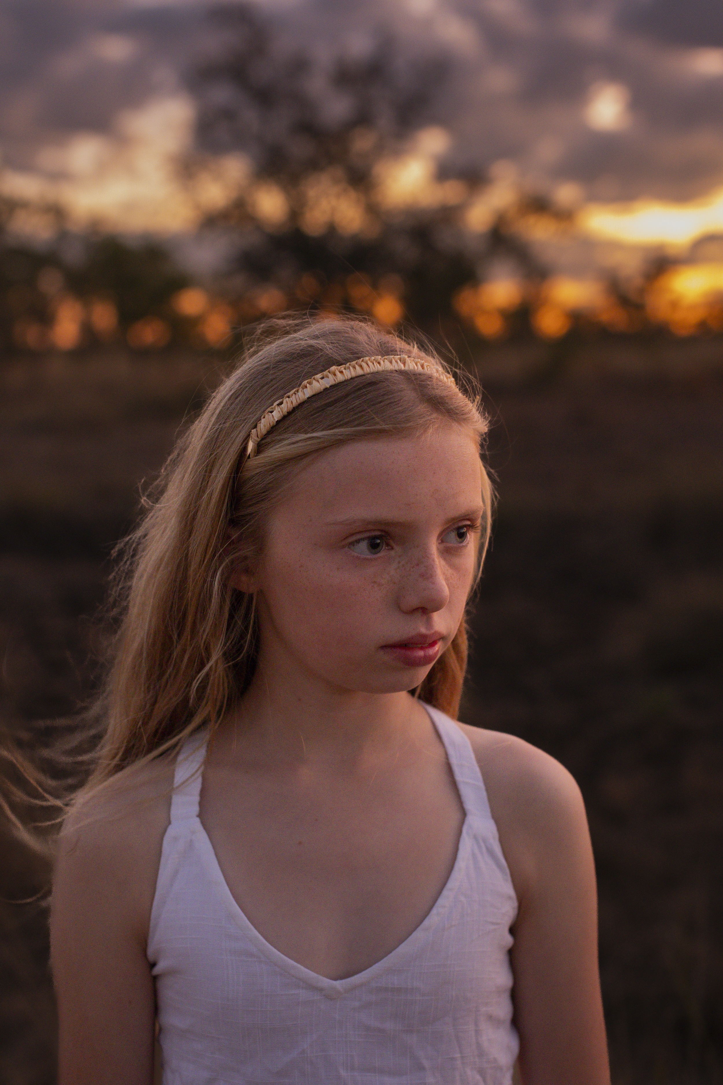 Young girl with freckles and blonde hair wearing a white sleeveless top and headband, looking thoughtfully to the side at sunset.