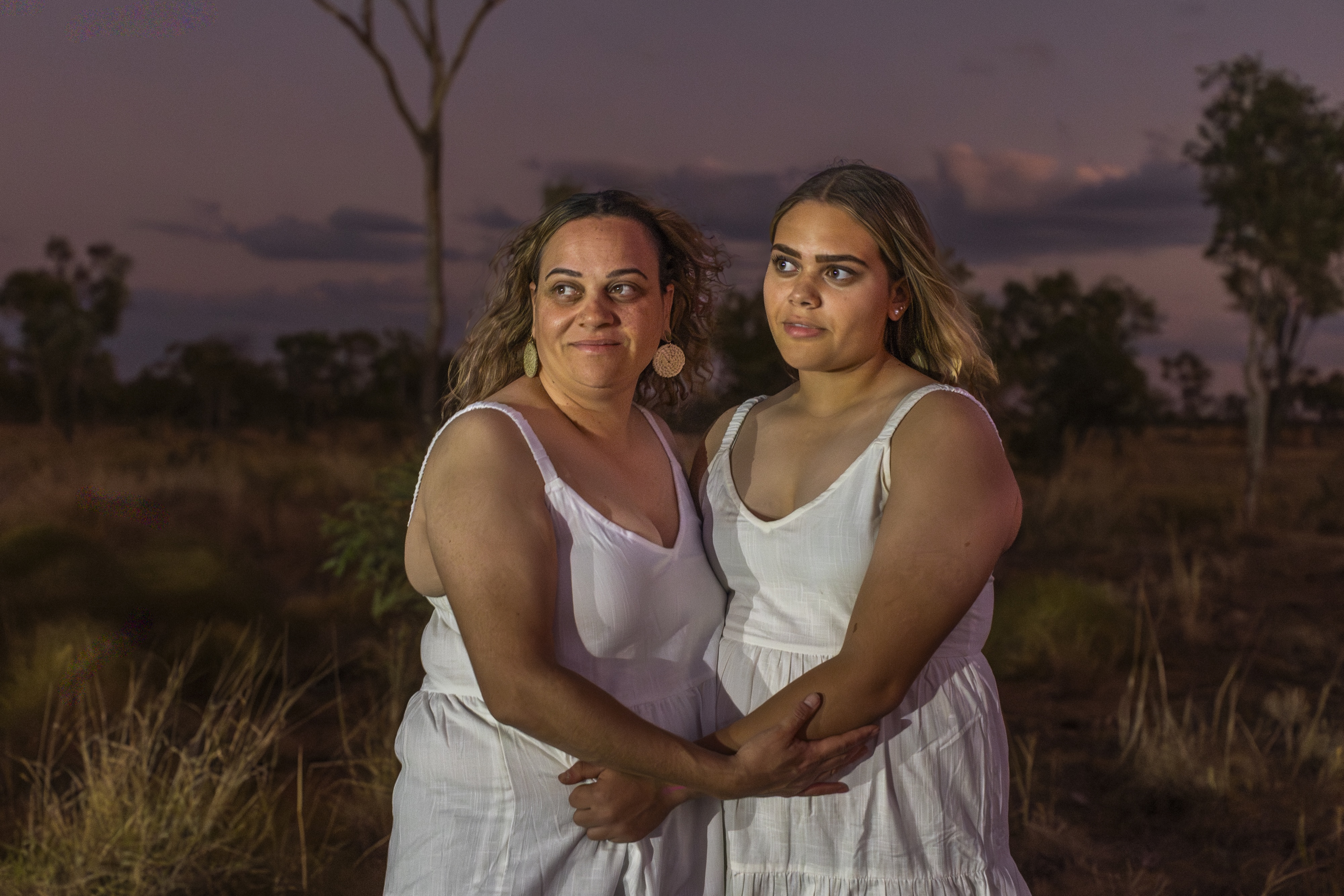 Two women in white dresses embracing outdoors at dusk with trees and grass in the background.
