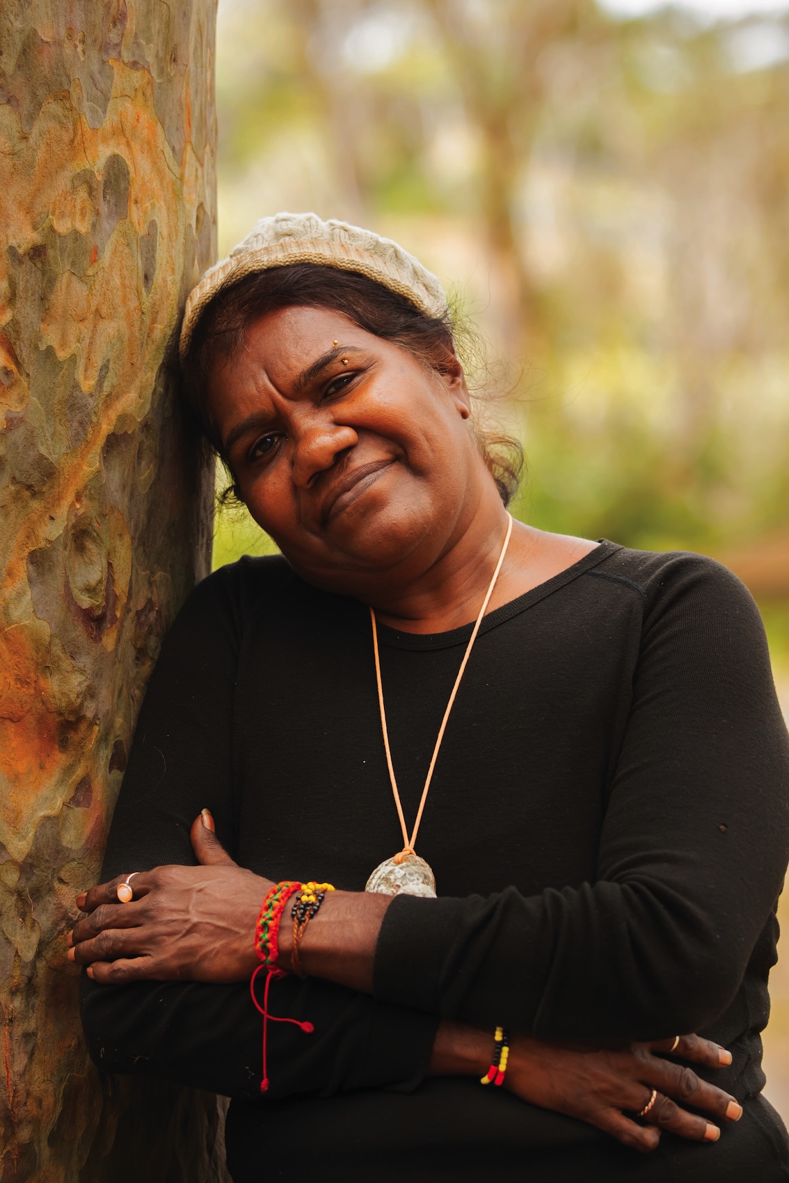 Woman wearing a black long-sleeve shirt and knit cap, leaning against a tree with arms crossed, outdoors.