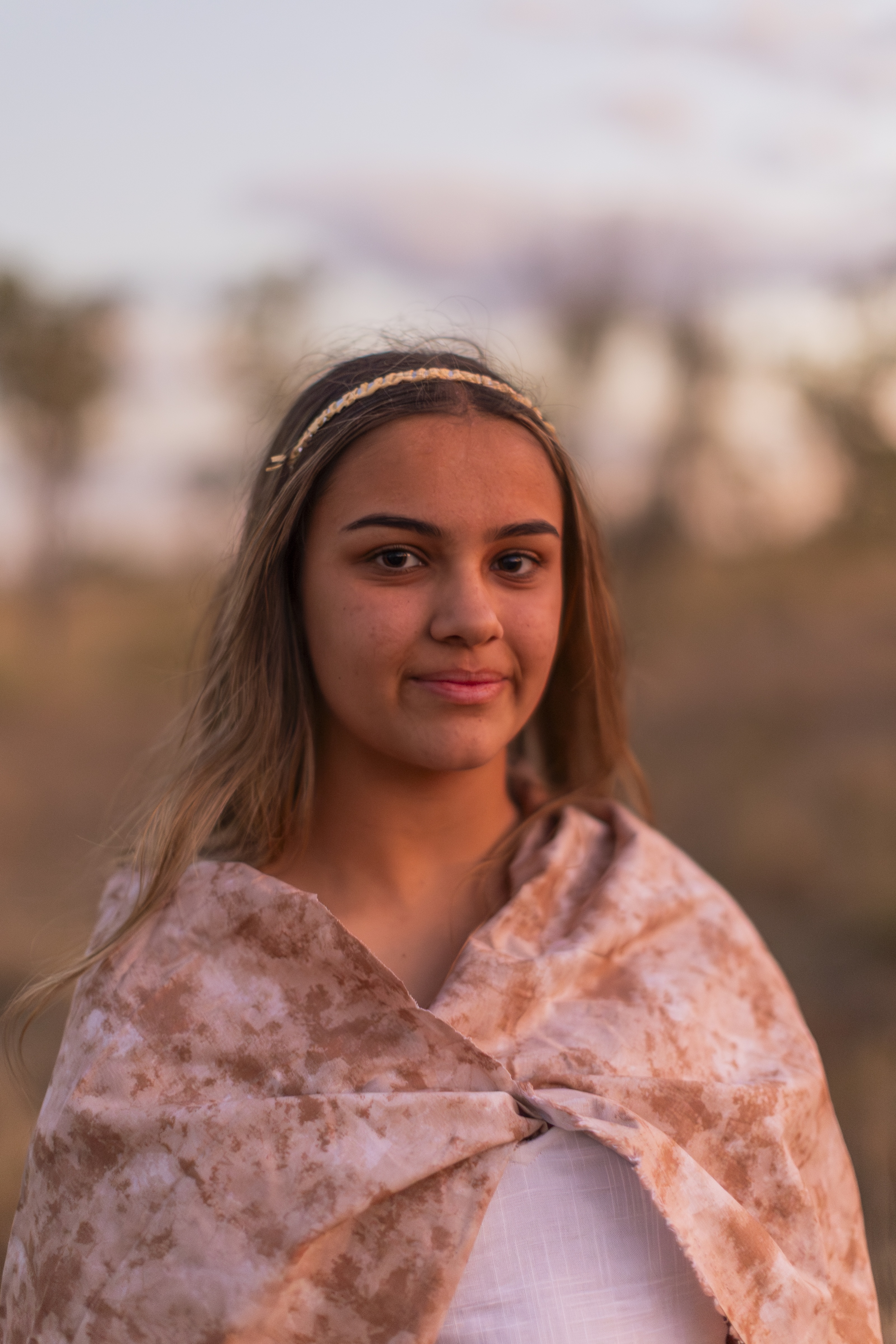 Young woman with a braided headband and light brown patterned shawl, standing outdoors at sunset.