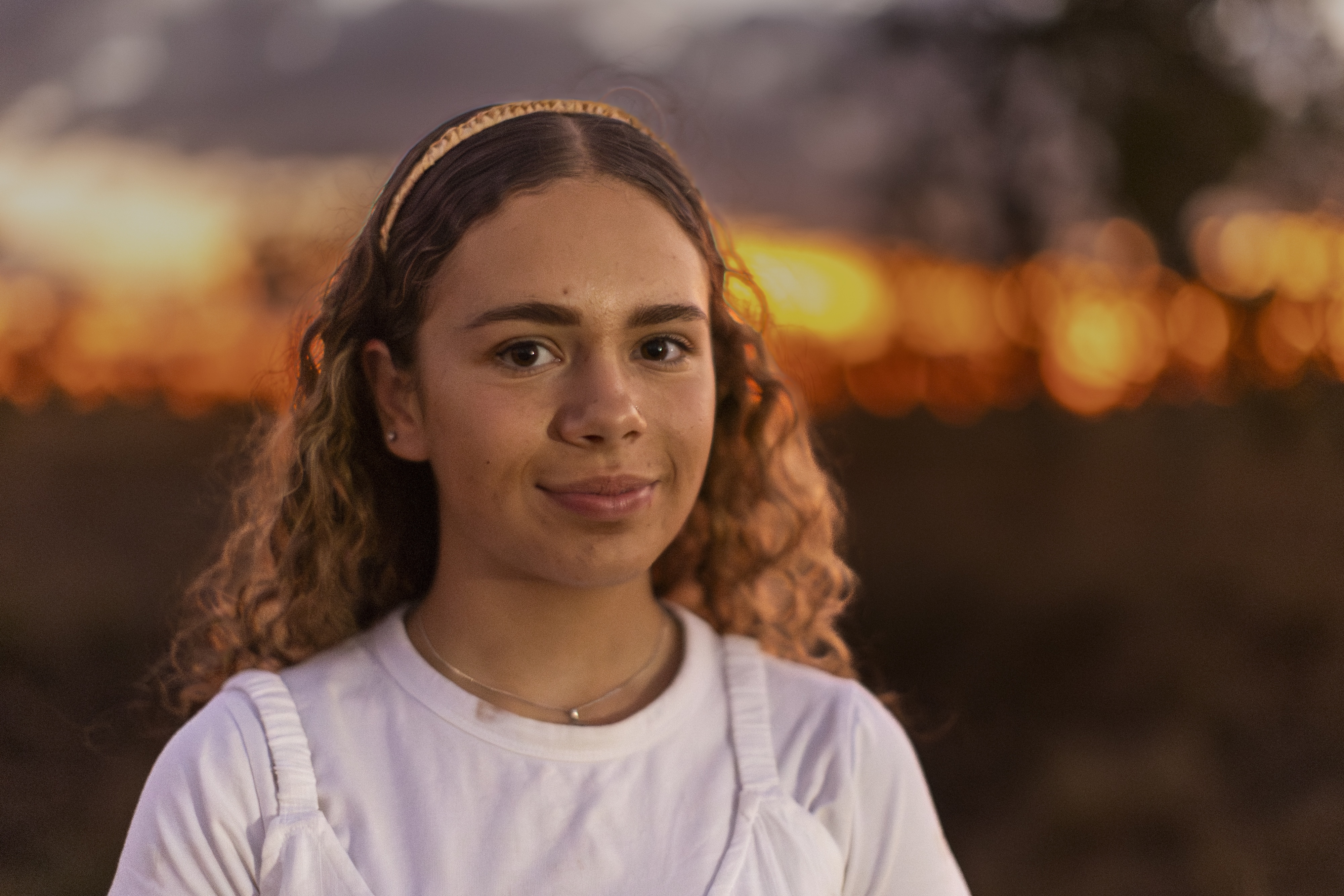 Young woman with curly hair wearing a white top and headband, smiling softly at sunset.