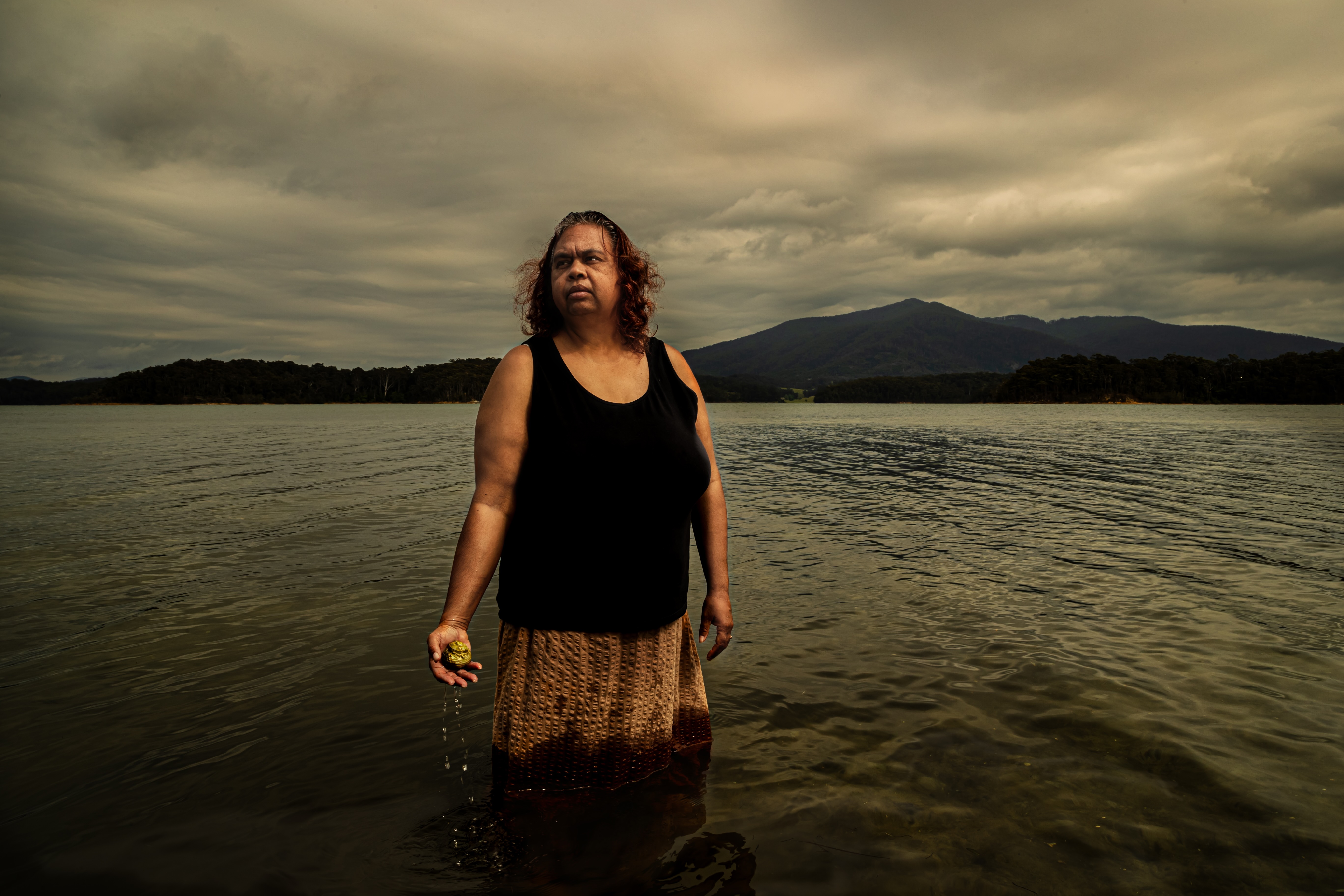 Woman standing waist-deep in water holding a dripping yellow object, with a cloudy sky and forested mountains in the background.