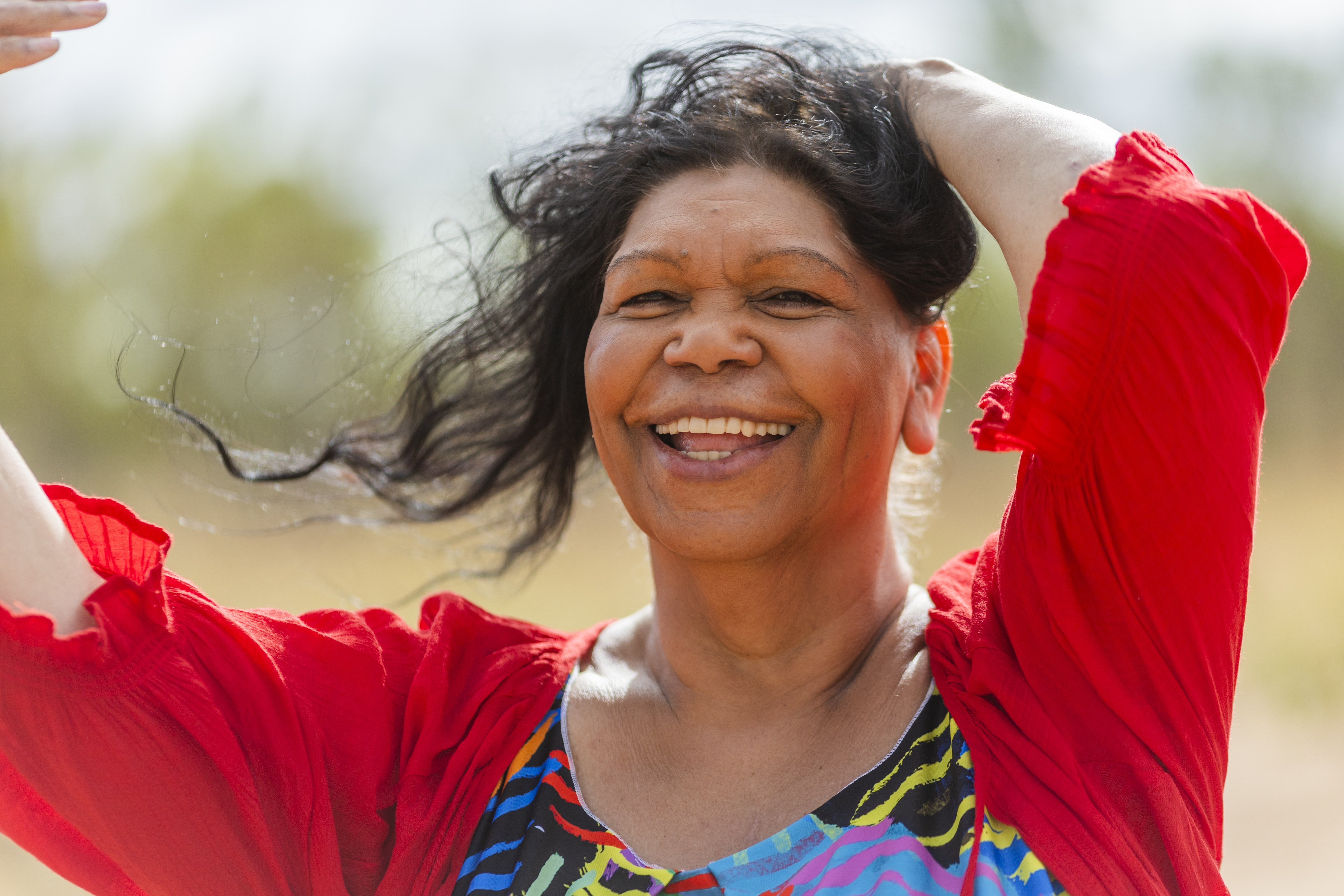 Smiling woman with flowing black hair wearing a colorful dress and a red shawl outdoors.