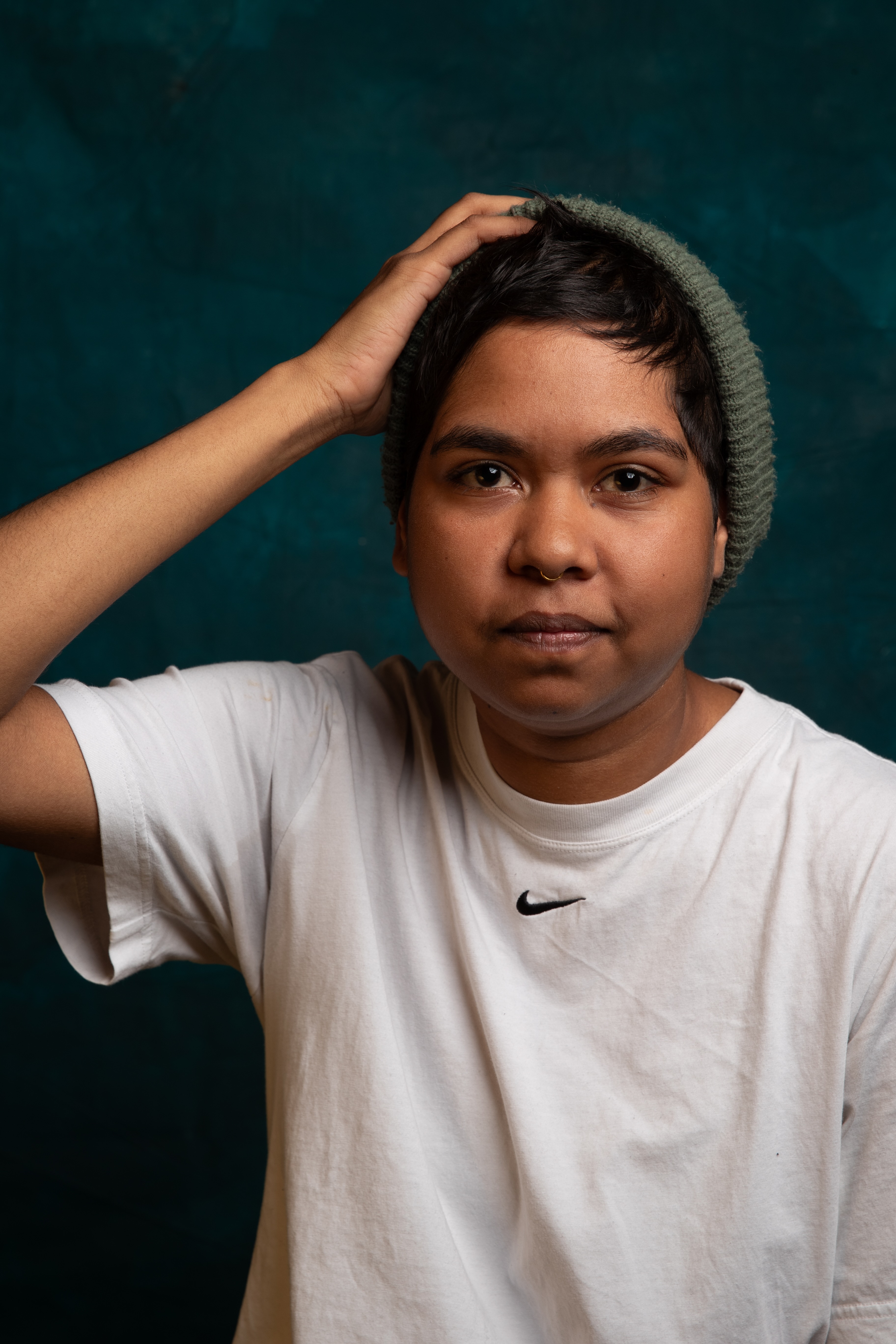 Person wearing a green knit beanie and white Nike t-shirt, looking directly at the camera with one hand on their head.