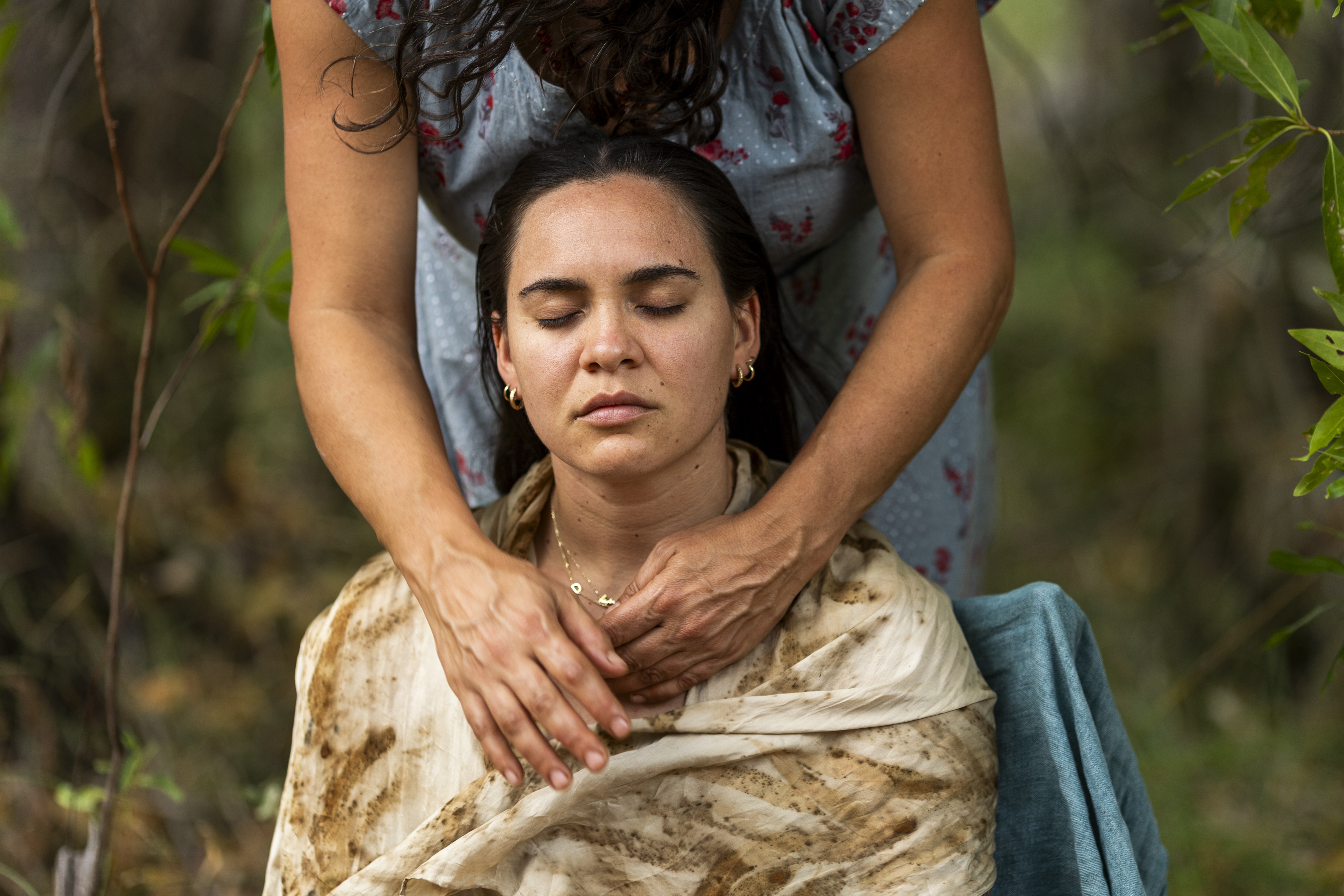 A woman with closed eyes wrapped in a dirt-stained cloth receives a neck massage from a standing person in a grey dress with red flowers, outdoors.