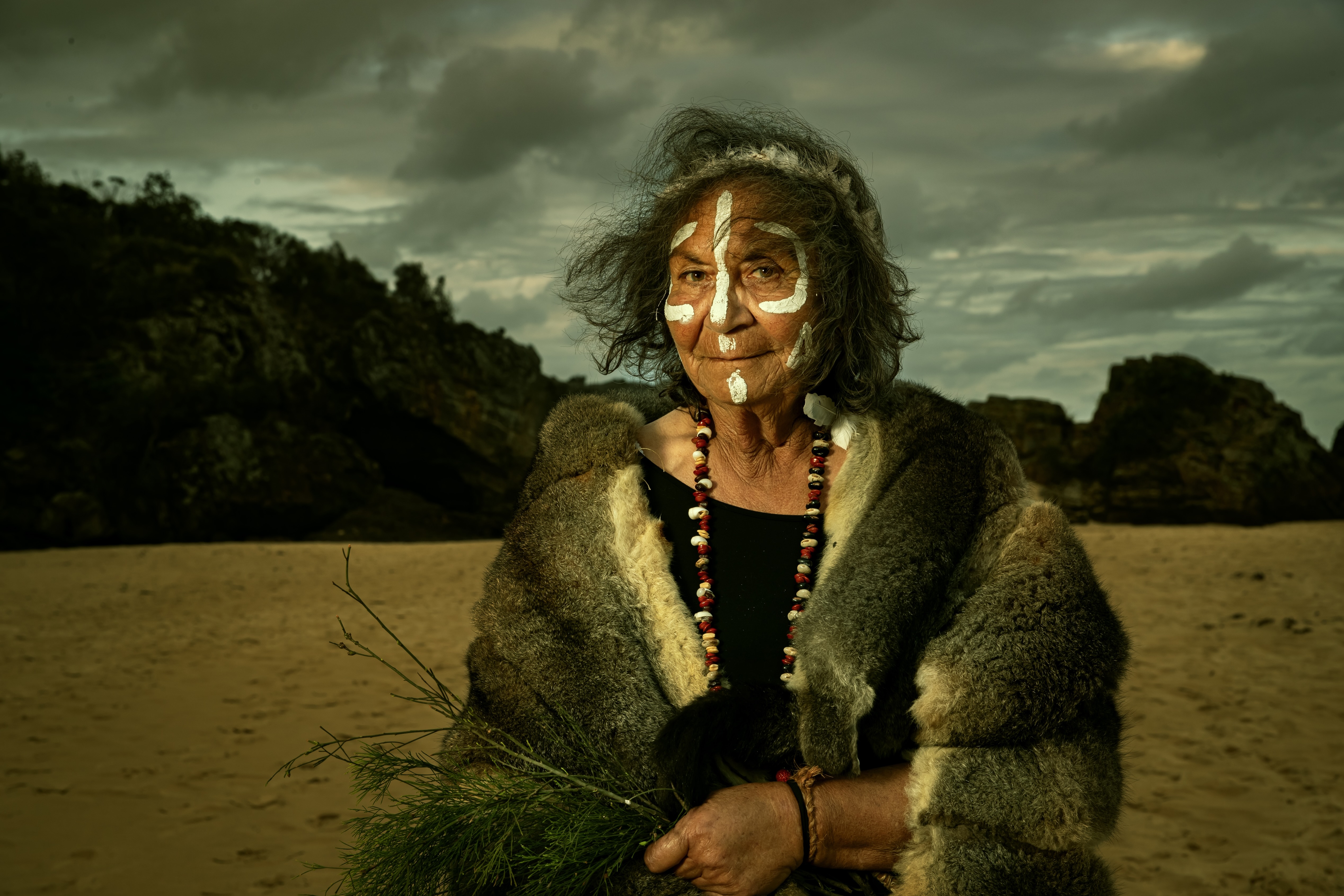 Elderly woman with traditional white face paint and a beaded necklace, wrapped in a fur cloak, standing on a sandy beach with rocky cliffs and a cloudy sky in the background.