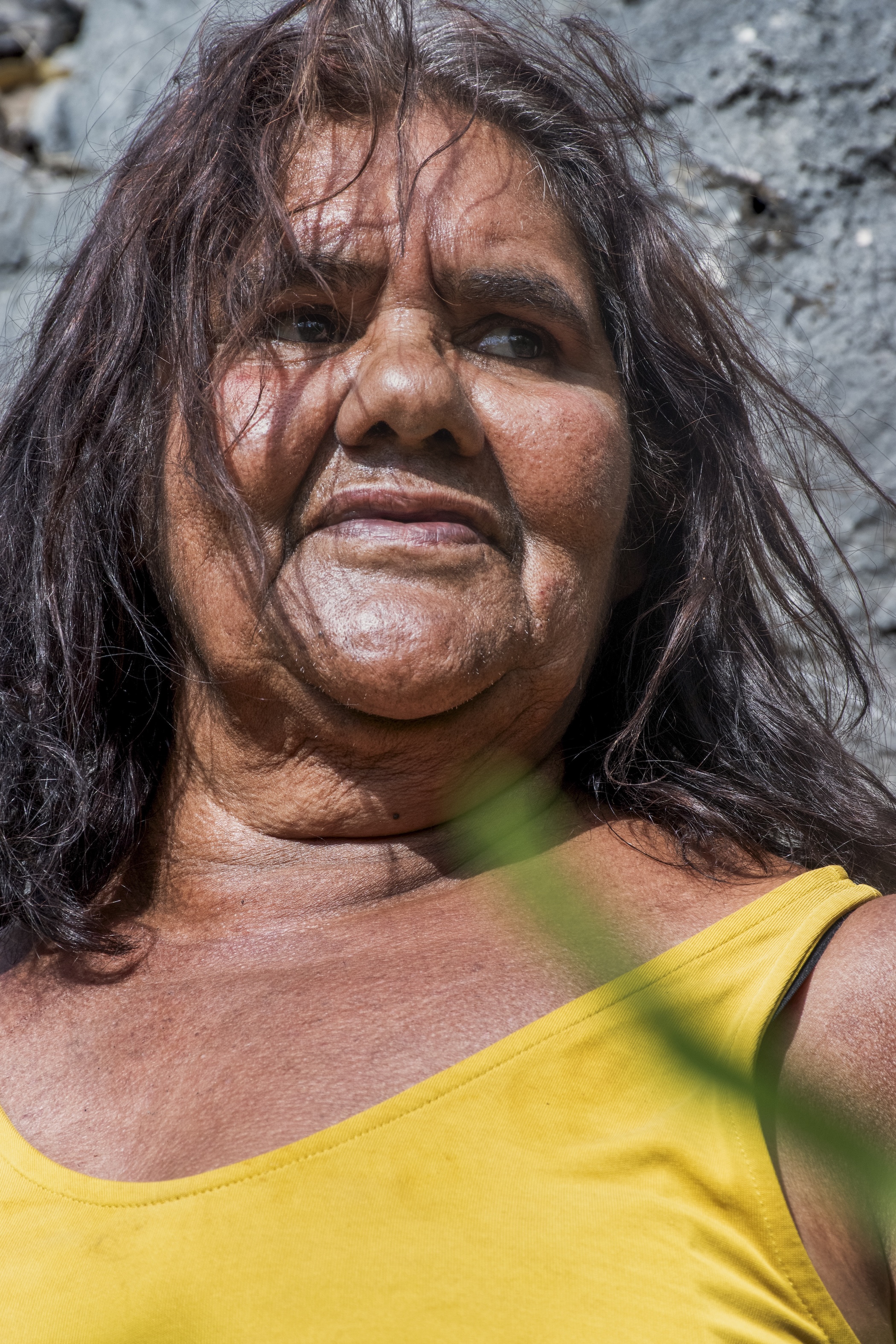Close-up of a woman with long dark hair wearing a yellow sleeveless top, looking slightly to the side against a textured gray wall.