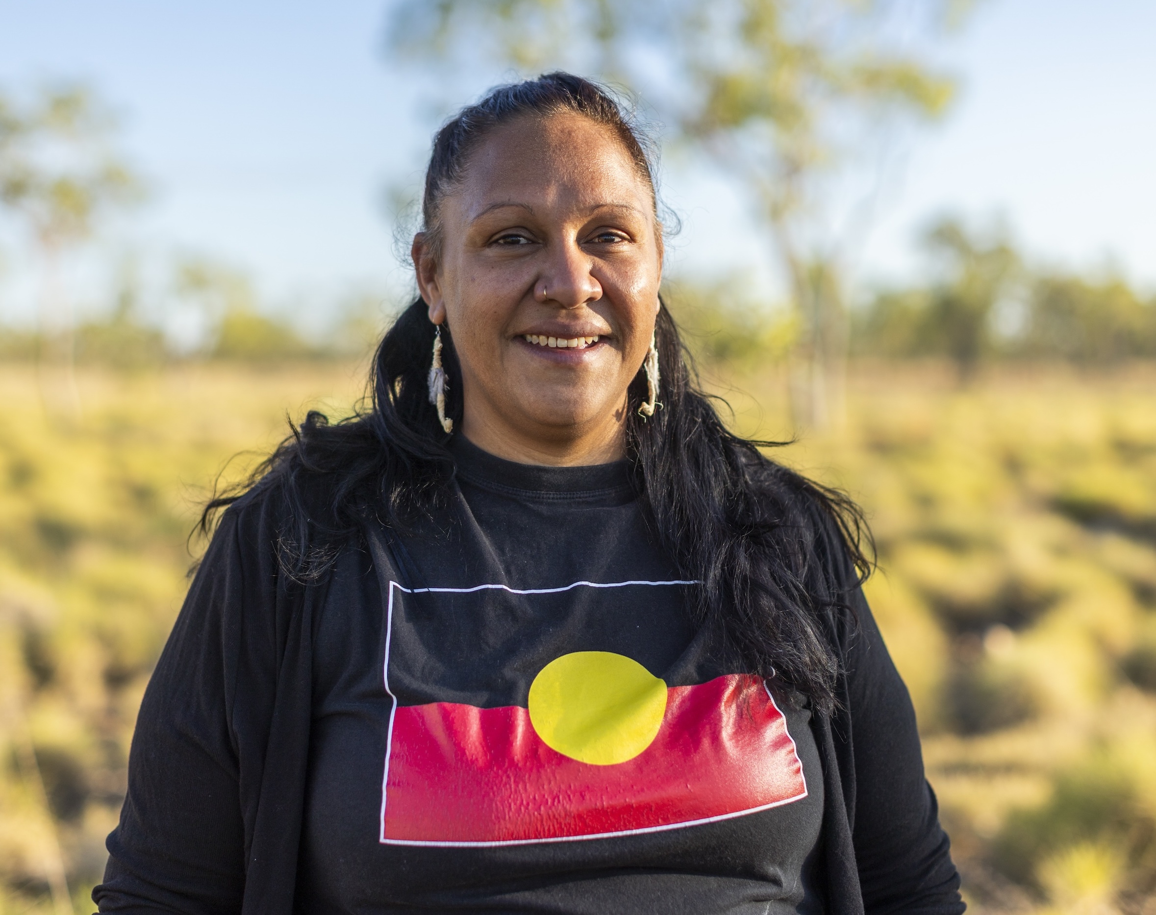 Woman outdoors wearing a black shirt with the Aboriginal flag, smiling with trees and grass in the background.