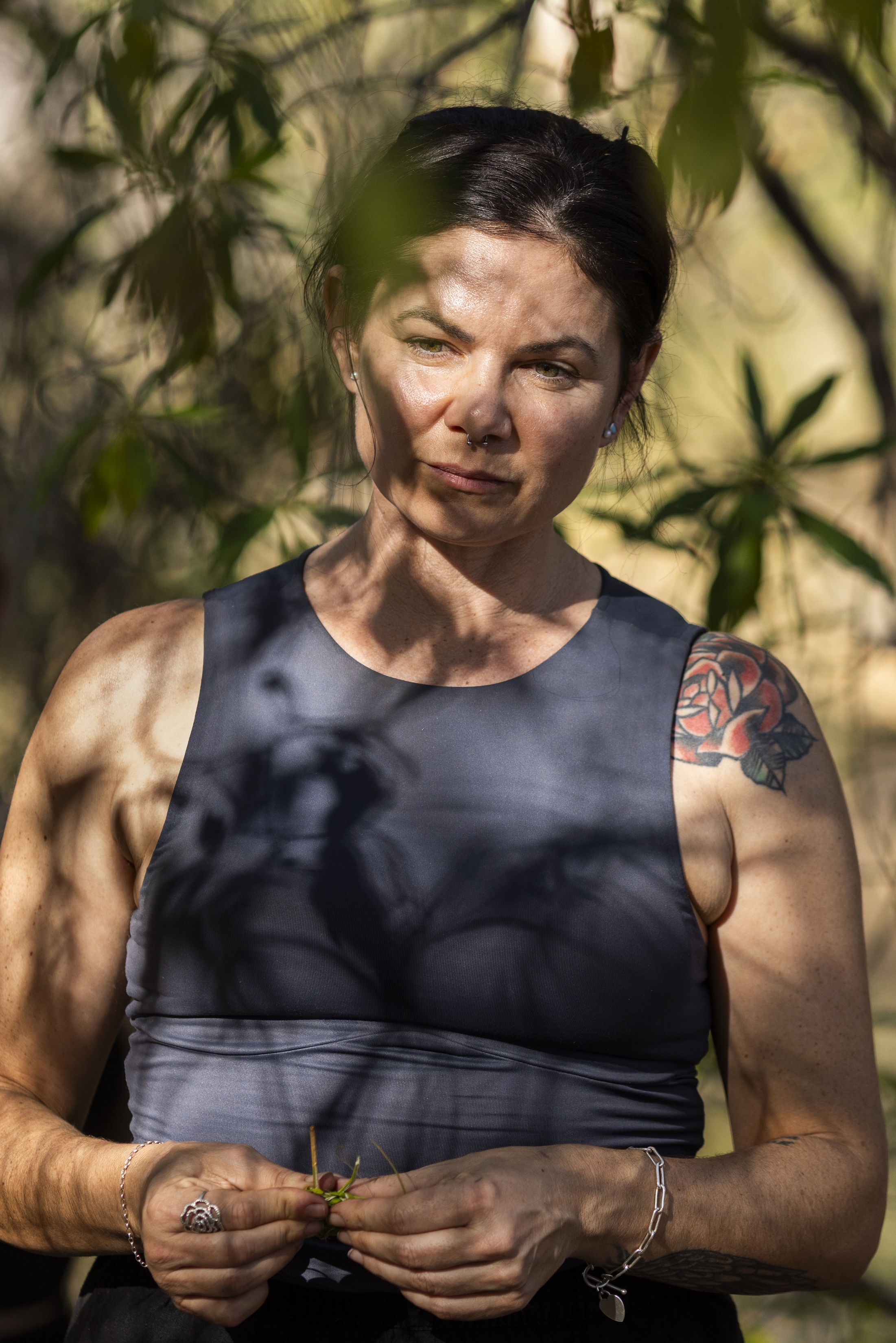 Woman with a septum piercing and floral tattoo on her shoulder, wearing a sleeveless top, standing outdoors among trees with shadows cast on her face and body.