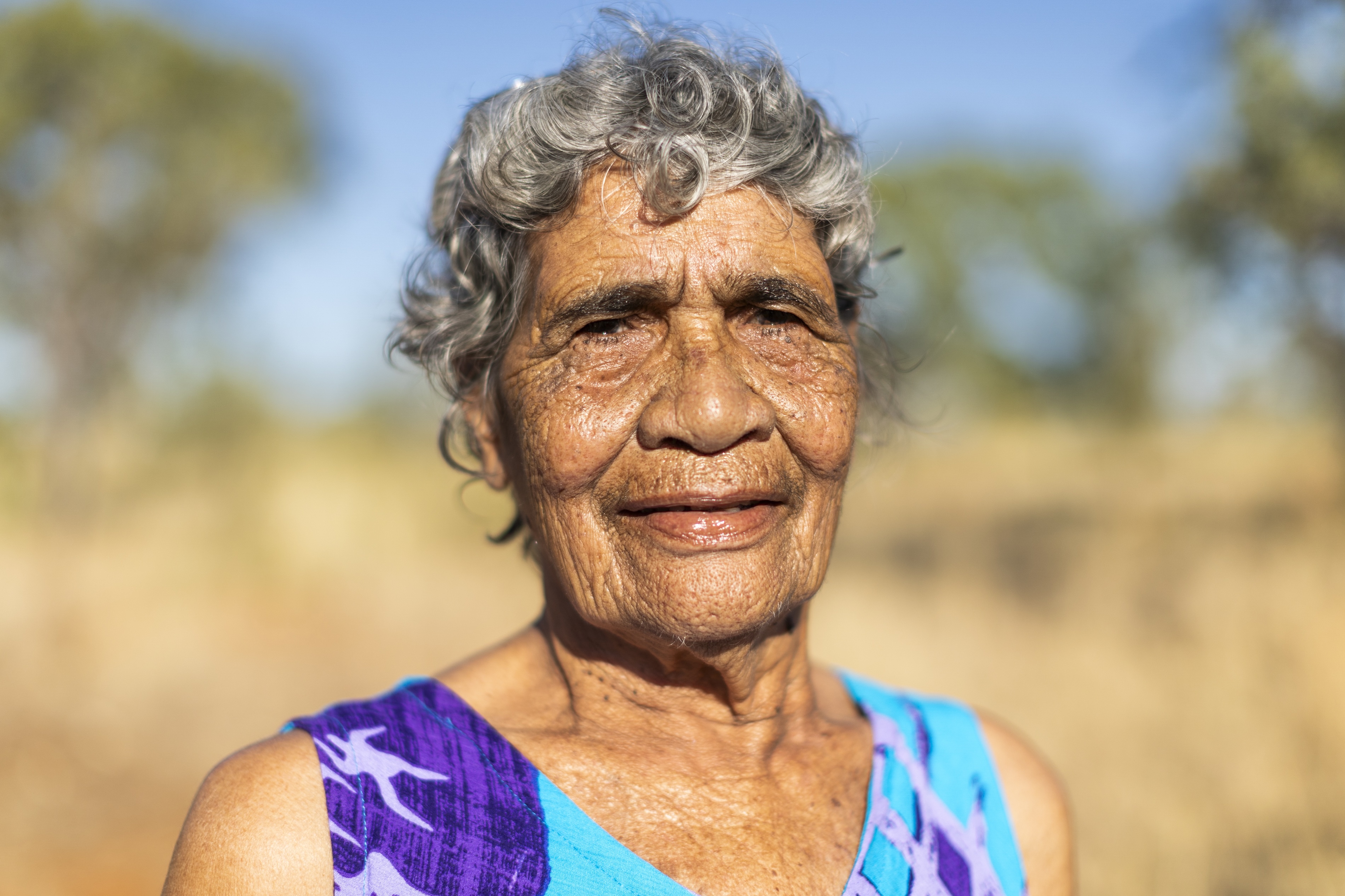 Portrait of an elderly woman with gray curly hair wearing a colorful blue and purple sleeveless top, standing outdoors with a blurred natural background.