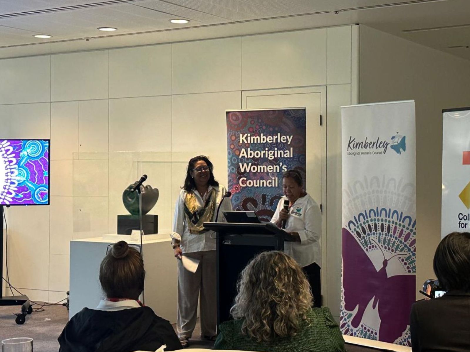 Janine Dureau and Selena O’Meara standing at lectern, talking to crowd.
