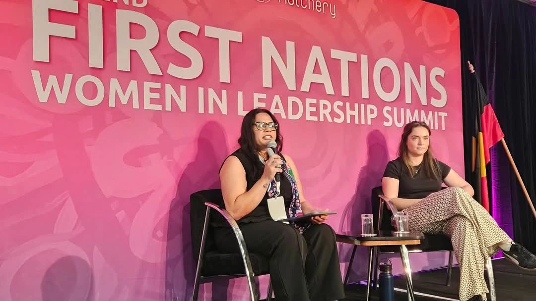 A photograph of a panel discussion at The 2nd First Nations Women in Leadership Summit. Two women are seated on stage in front of a large pink backdrop that reads: “FIRST NATIONS WOMEN IN LEADERSHIP SUMMIT” The woman on the left is speaking into a microphone, holding a notepad. She wears glasses and a black outfit. The woman on the right sits with her legs crossed, listening attentively, dressed casually in patterned pants and a black shirt. Behind them is an Aboriginal flag standing vertically on the right side of the stage. A glass of water and water bottle sit on the table between them.