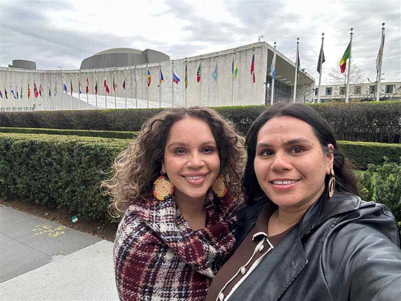 Two women, Banok Rind and Terri Reid, are taking a selfie in front of a building. They are outdoors, smiling and dressed in jackets and scarves. The setting includes a clear sky with some clouds above.