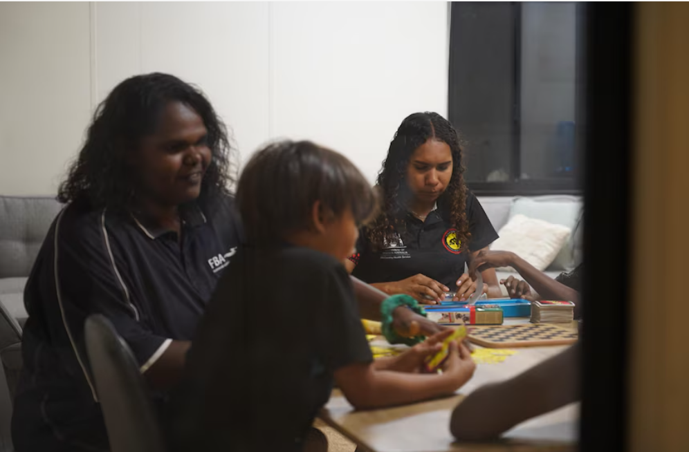 A photograph showing an Aboriginal woman and children engaged in an activity around a table in what appears to be an indoor setting. They are wearing dark clothing and appear to be working with materials or games on the table. The image is taken through a doorway, creating a candid, documentary-style perspective of the interaction.
