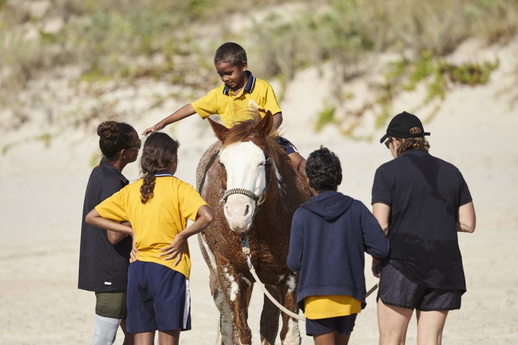 A young First Nations child in a yellow school uniform sits atop a brown and white horse on a sandy beach, surrounded by a small group of children and an adult. The children look on with interest as the horse is led along the sand, with coastal dunes and vegetation visible in the background.