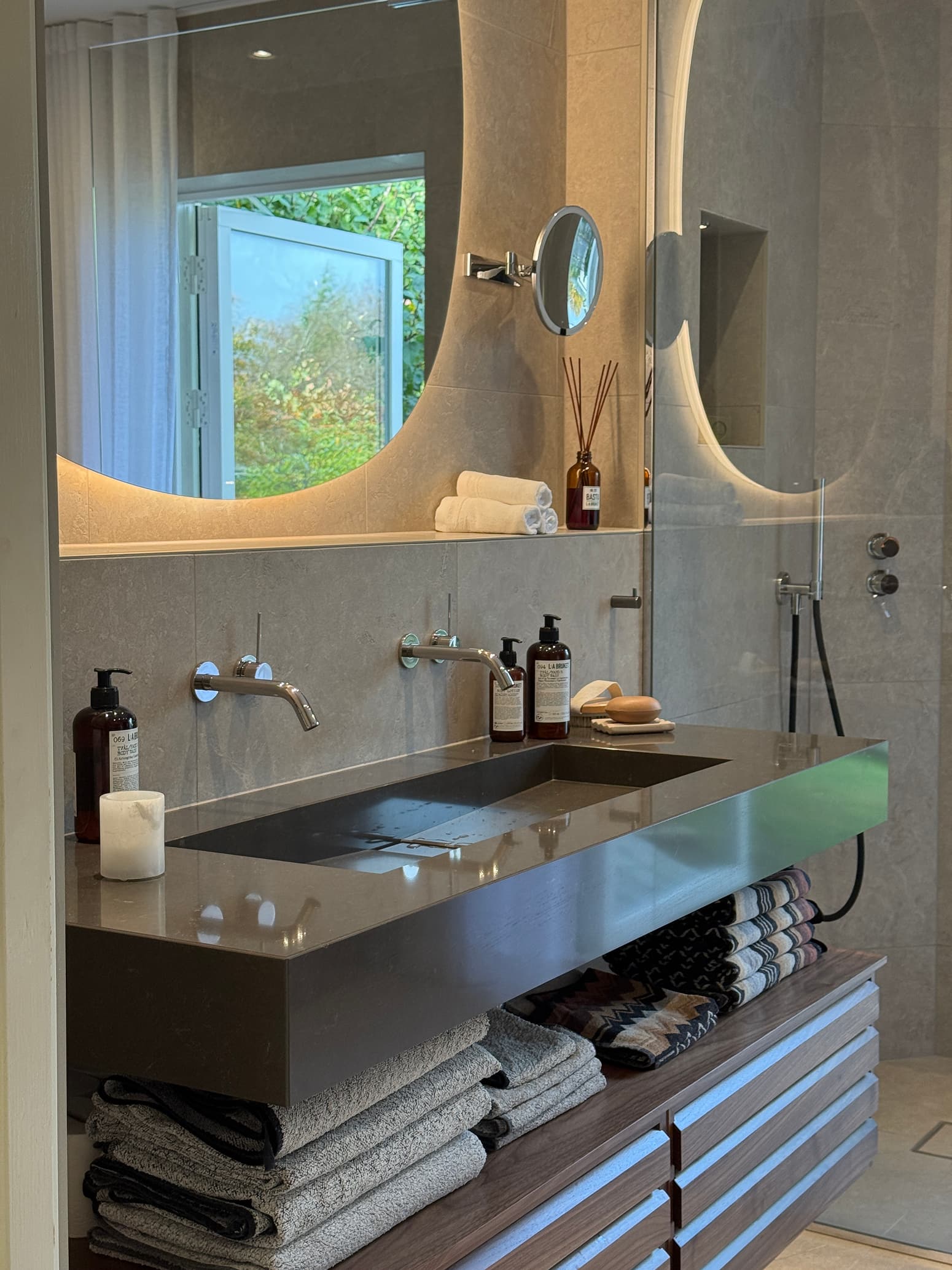 Modernt badrum med dubbel handfatslösning i sten, väggmonterade kromblandare och bakbelyst spegel ovanför.