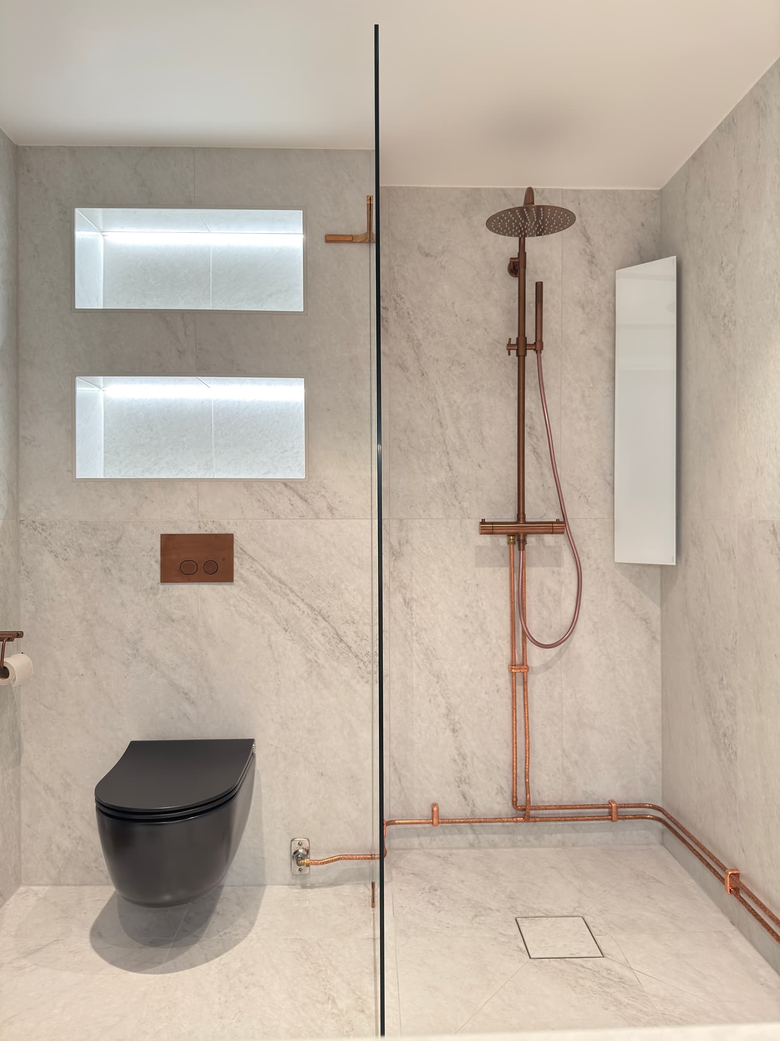 Minimalistiskt badrum med svart toalett, synliga kopparrör och regndusch mot ljus marmorinspirerad vägg.