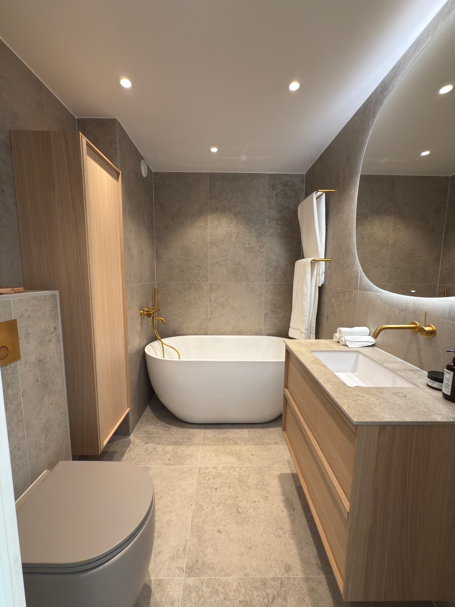 Elegant badrum med fristående badkar, beige kakel, trädetaljer och mässingsfärgade blandare.