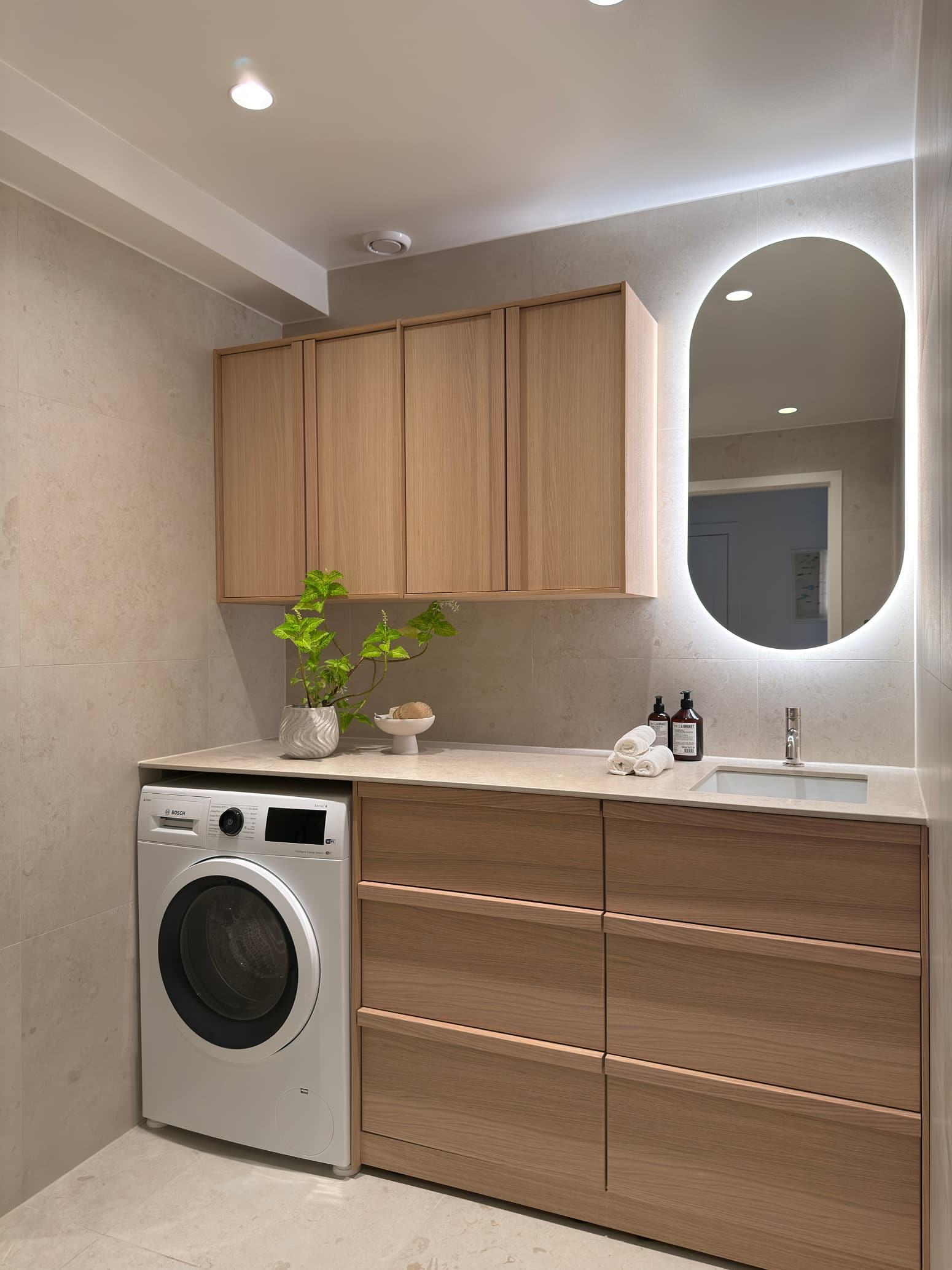 Ljust badrum med integrerad tvättmaskin, träskåp, bakbelyst spegel och stilren inredning.