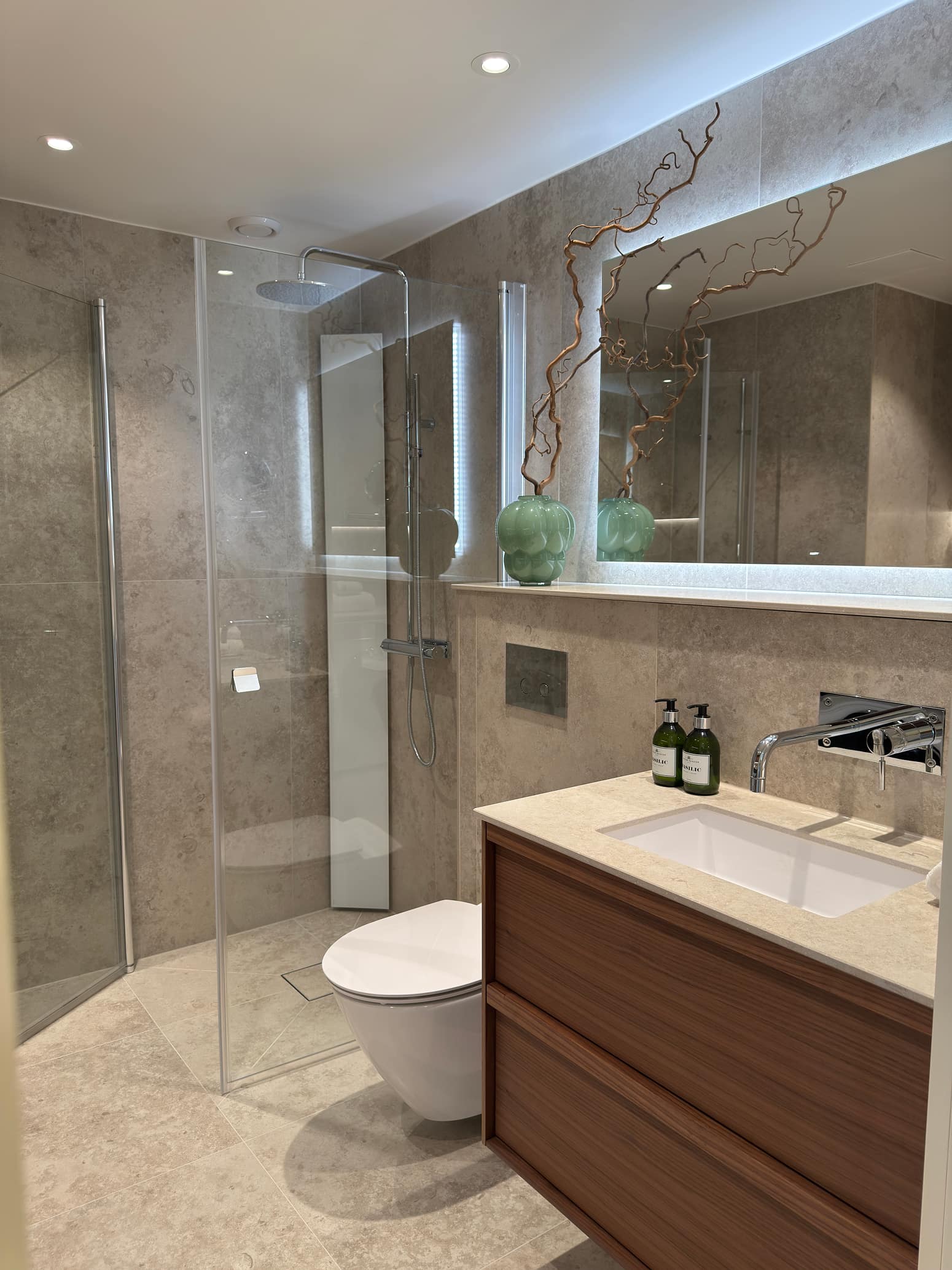 Modernt badrum med glasdusch, träkommod, steninspirerat kakel och dekorativa gröna vaser.