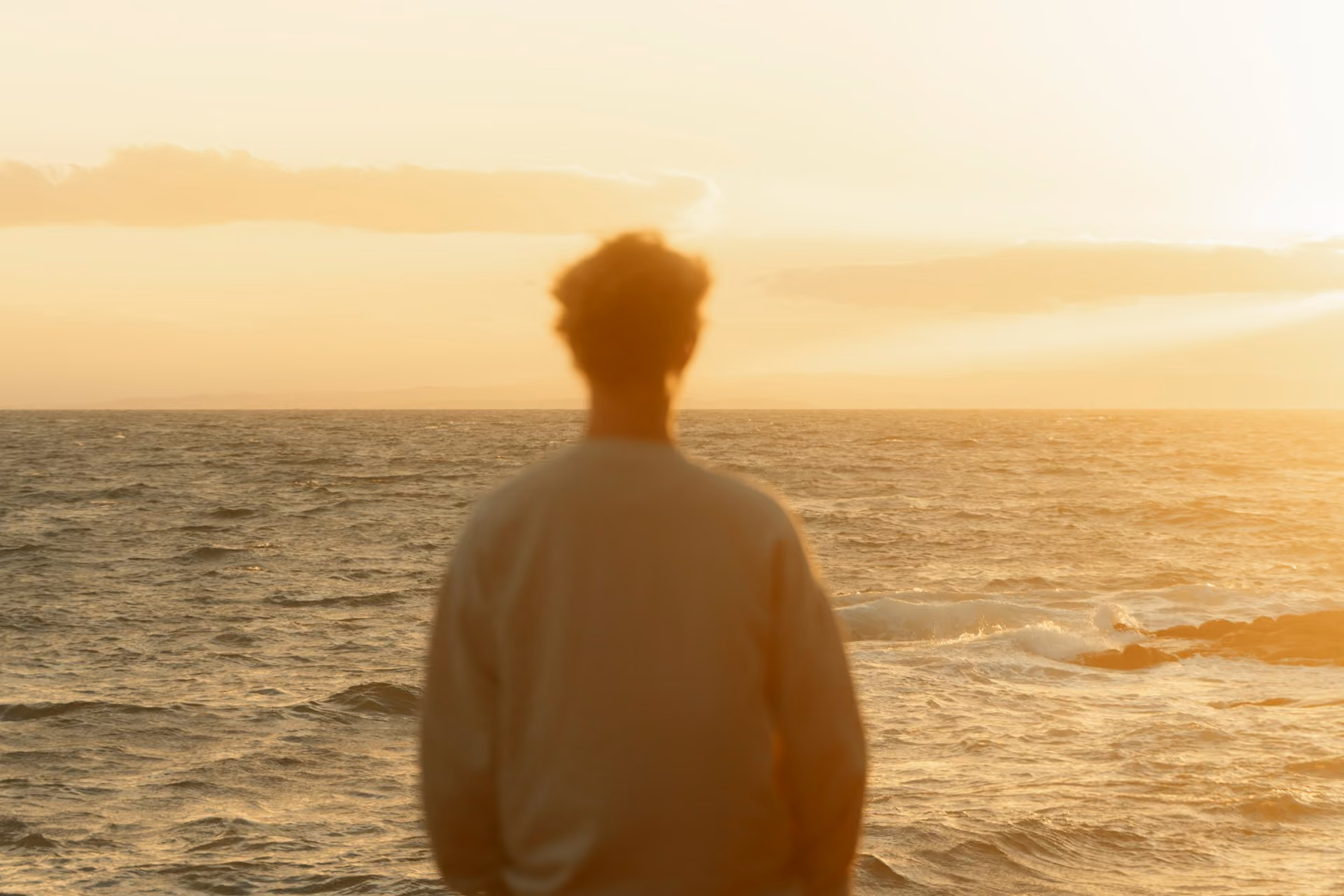 Person standing facing the ocean during sunset with warm orange light and waves visible on the water surface.