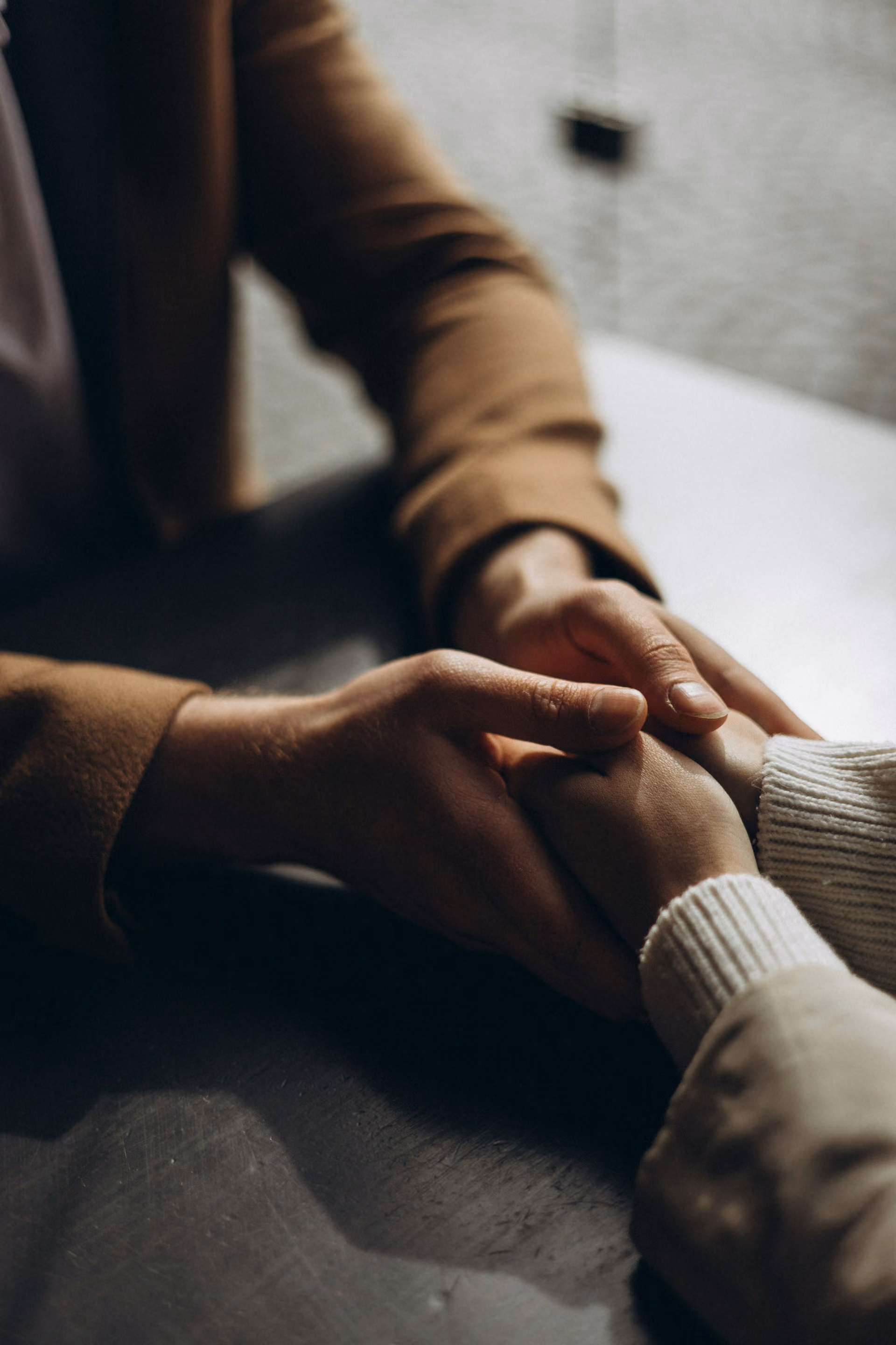 Two people sitting at a table holding hands in a comforting gesture, one wearing a brown jacket and the other a white sweater.