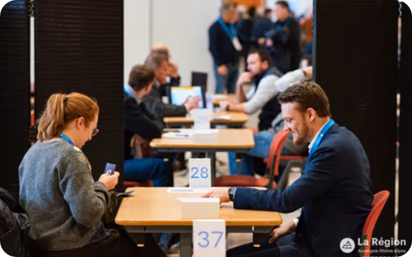 People seated across from each other at tables arranged in a row, participating in a professional networking or business event, with numbered table signs visible.