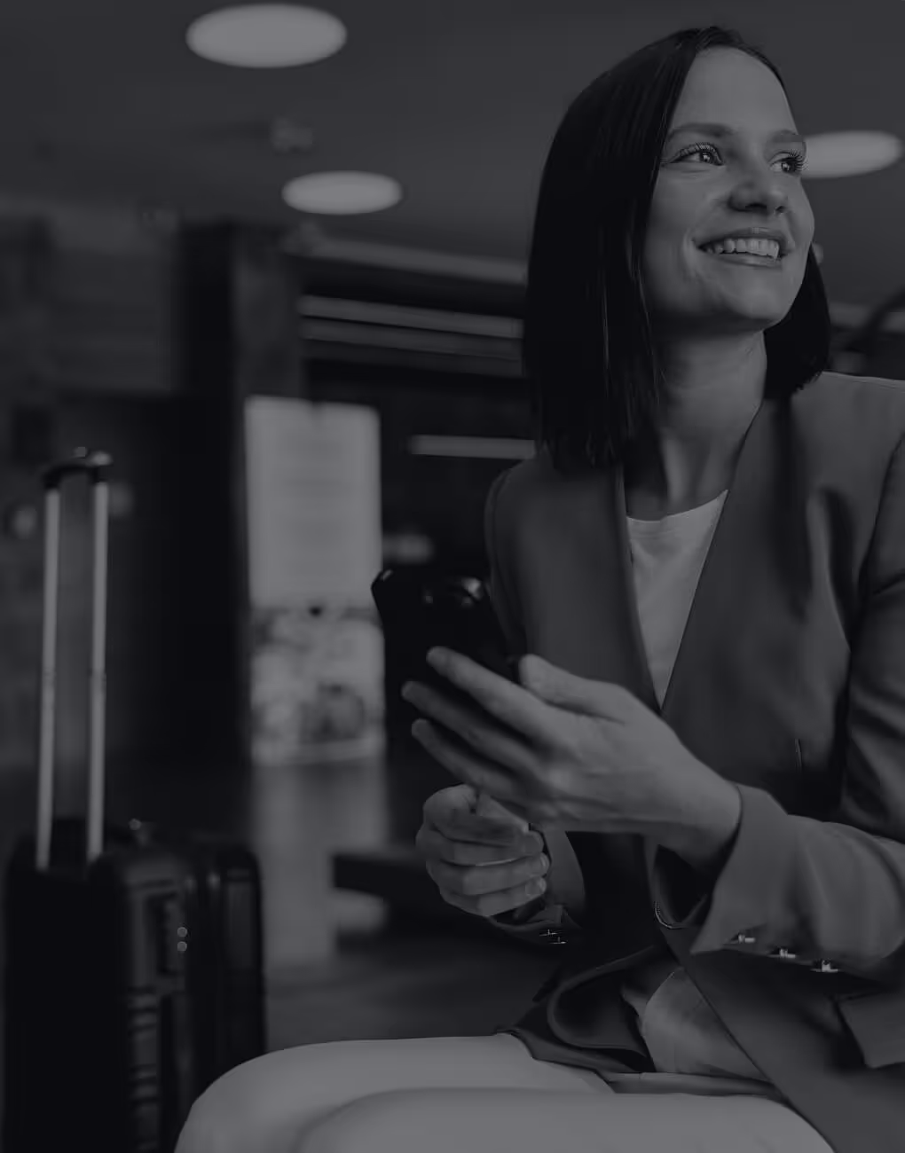 Smiling woman sitting with a suitcase, holding a smartphone in a modern indoor setting.