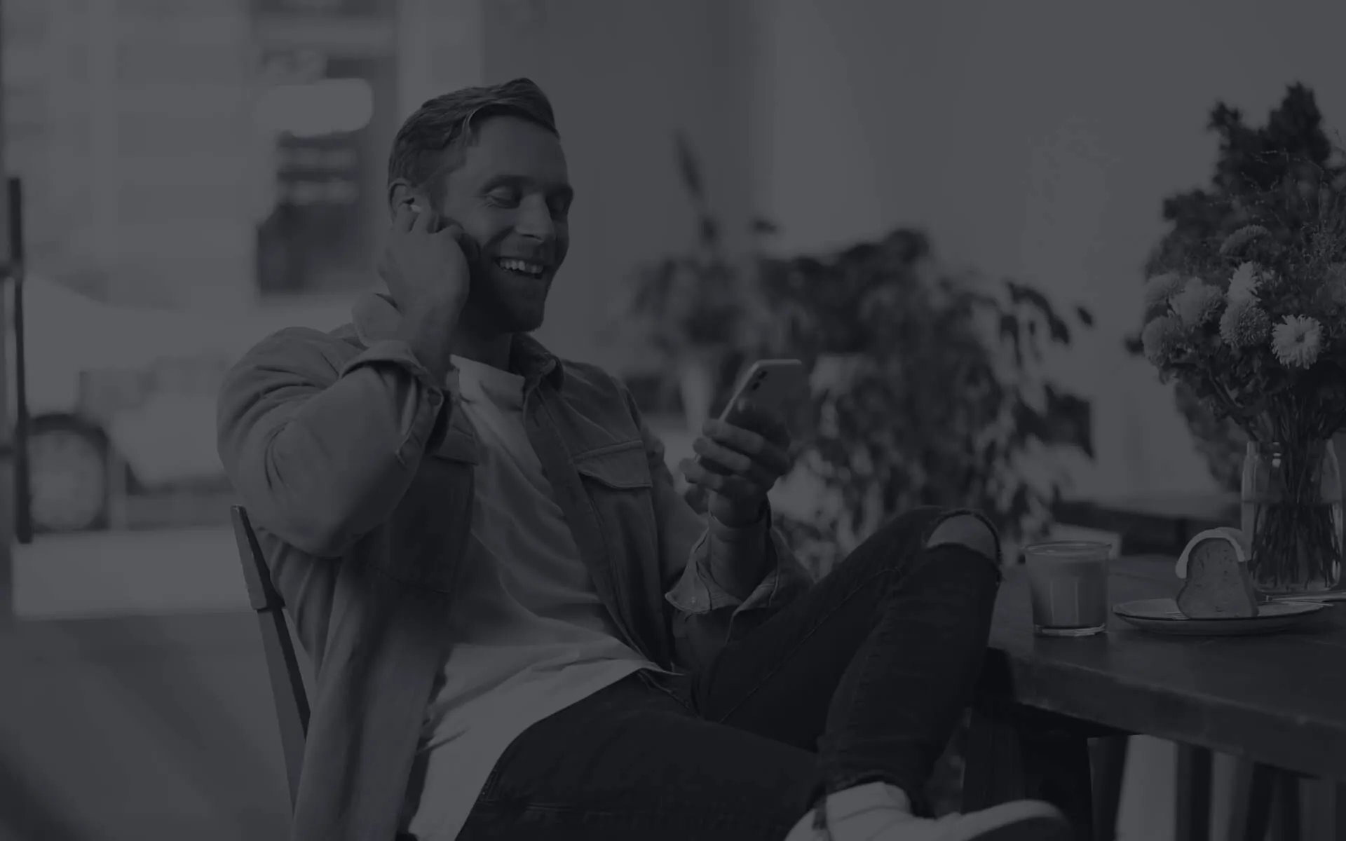 Smiling man sitting casually, adjusting a bluetooth earpiece while looking at his phone in his hand