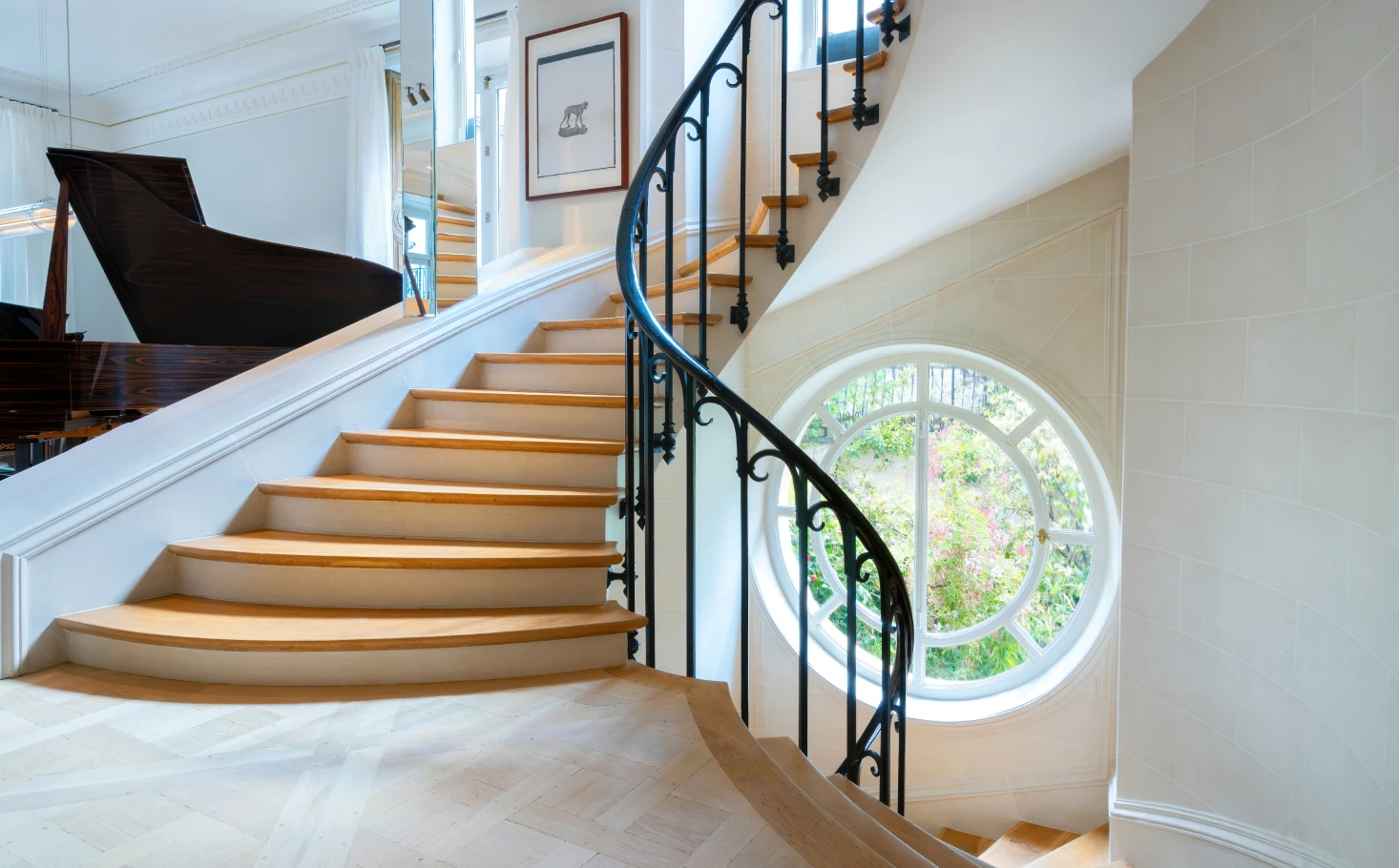 Elegant curved wooden staircase with black wrought iron railing and a large round window revealing greenery outside.