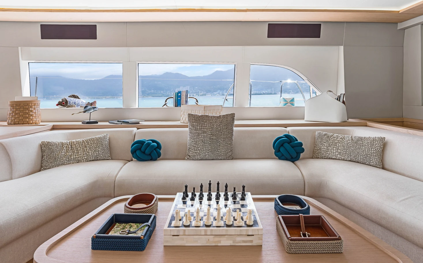 Modern yacht interior with a U-shaped beige sofa, decorative pillows, and a chessboard on a wooden table in front of large windows showing a sea view.