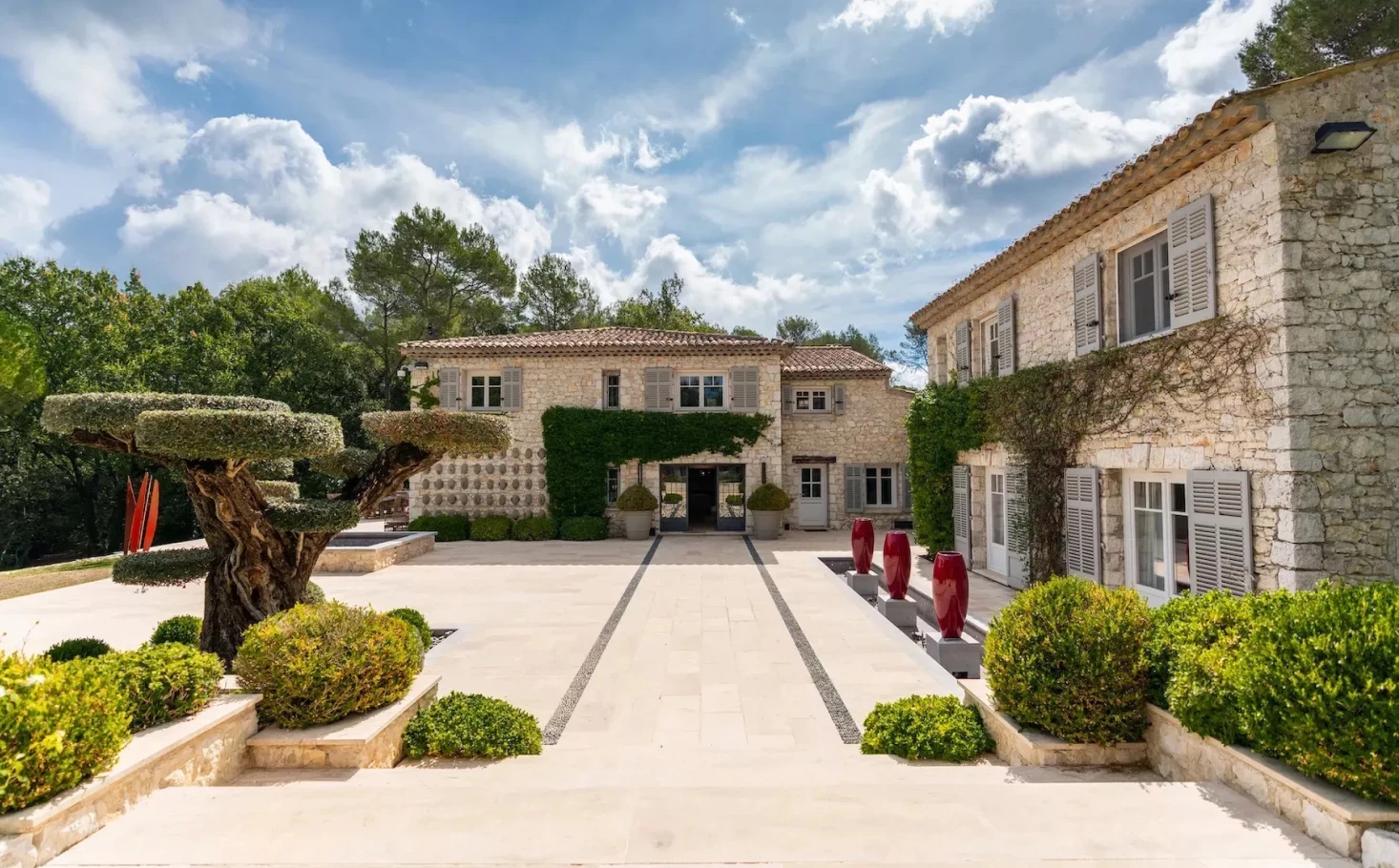A luxurious Mediterranean-style villa with stone facades, lush greenery, and a large driveway featuring neatly trimmed bushes and an elegant courtyard under a bright blue sky with clouds the valbonne