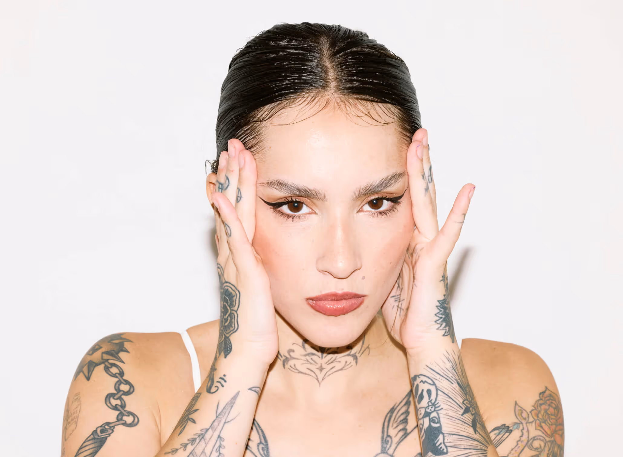 Close-up portrait of a woman with slicked-back hair, bold eyeliner, red lipstick, and multiple tattoos covering her arms, neck, and hands, holding her face.