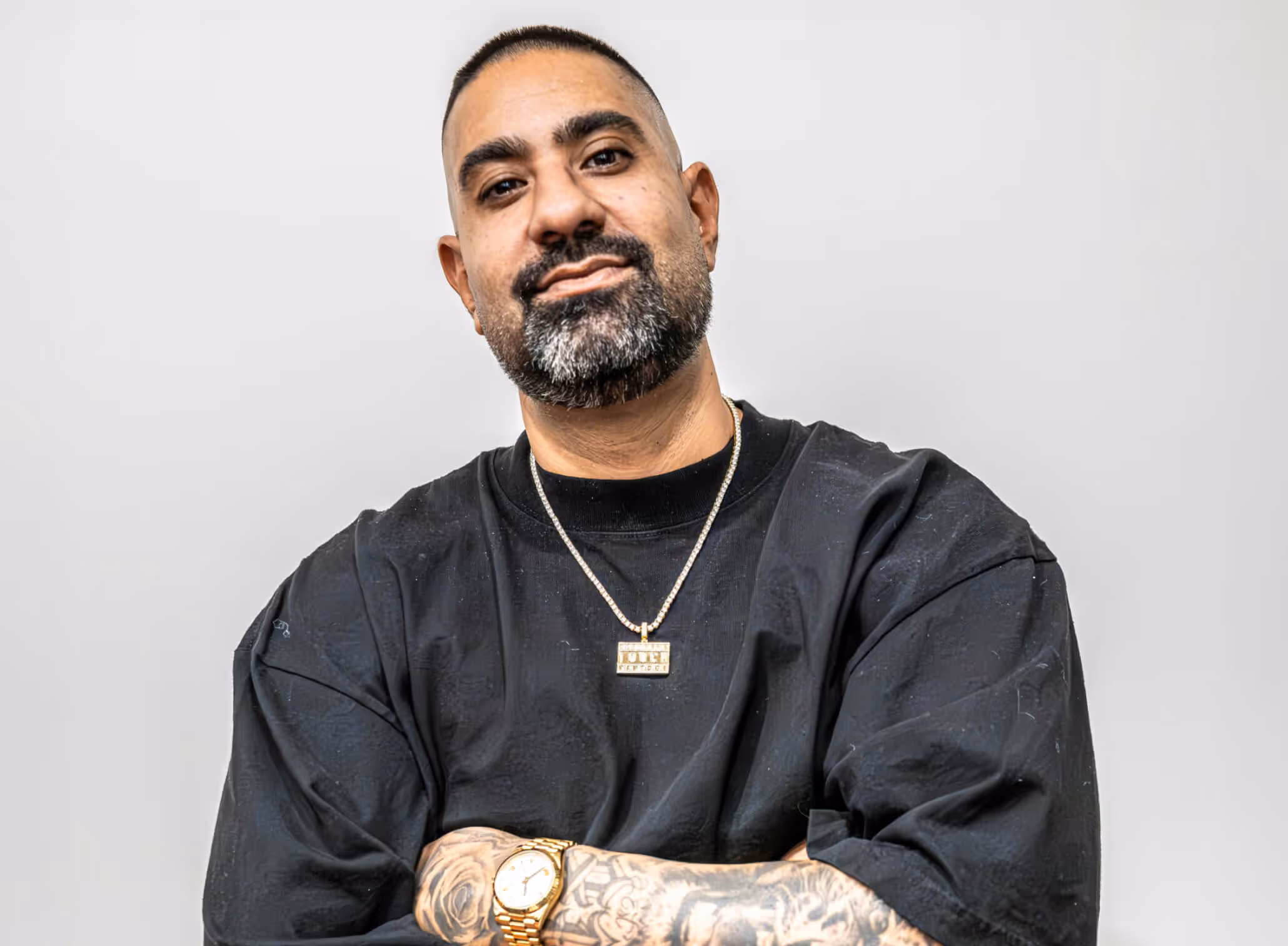 Man with a beard wearing a black shirt, gold watch, and a gold pendant necklace, standing against a plain light gray background with arms crossed.