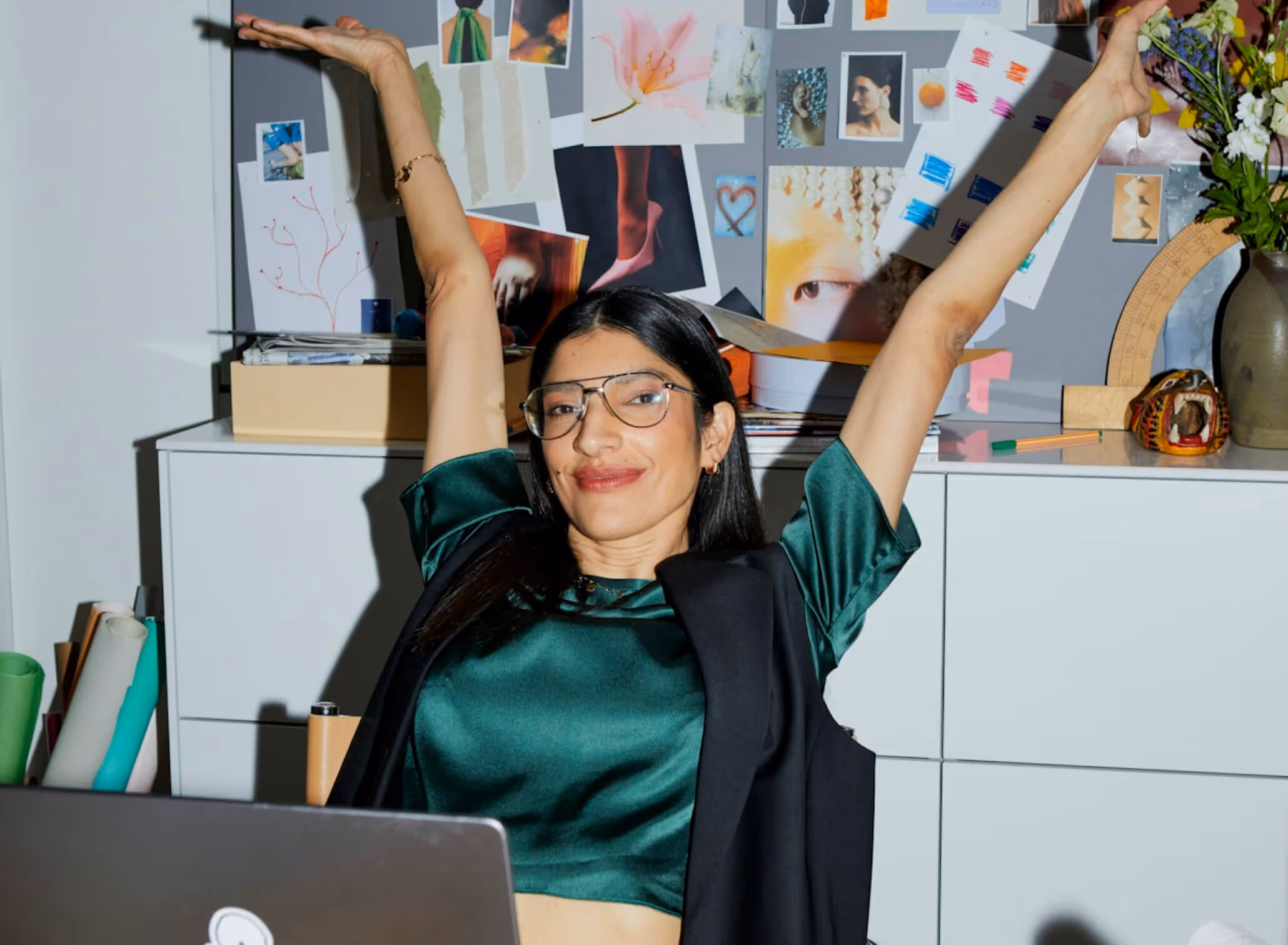 Smiling woman with glasses stretching arms up while sitting at a desk with a laptop and a collage of photos on the wall behind her.